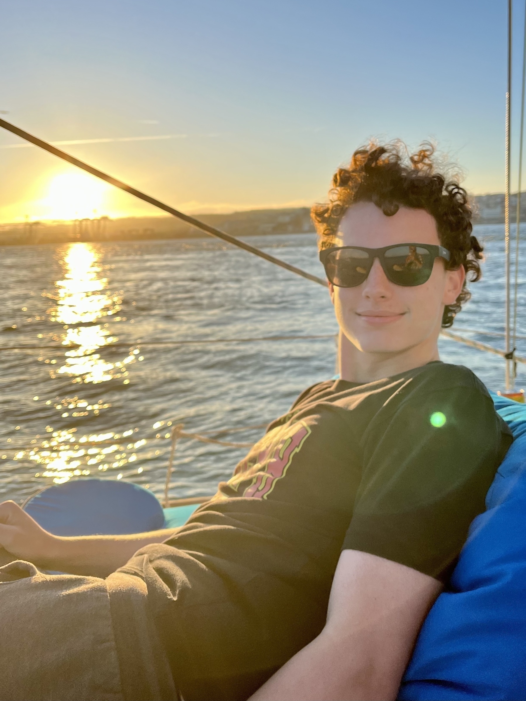 A person with sunglasses is sitting on a boat, relaxed, with a sunset and shimmering water in the background.