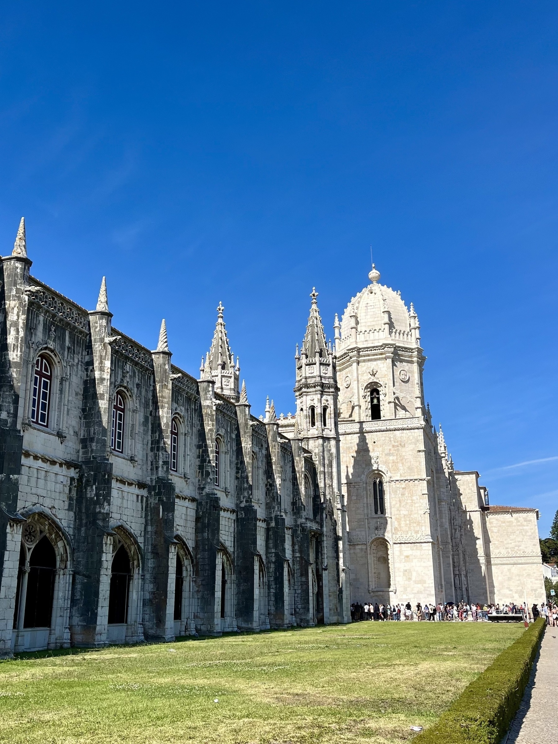 A Gothic monastery stands under a clear blue sky, with visitors queuing outside, set against a backdrop of well-kept green lawns.