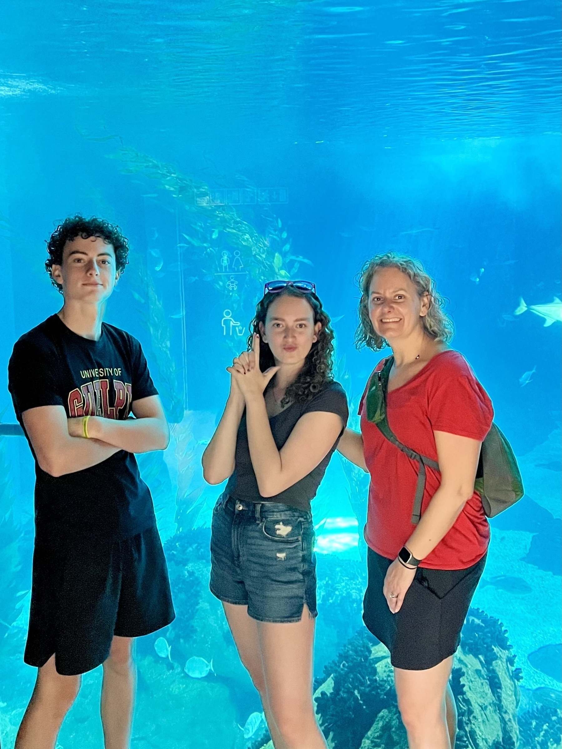 Three individuals pose in front of an aquarium with various fish and underwater flora, giving a sense of a leisure visit to a marine exhibit.