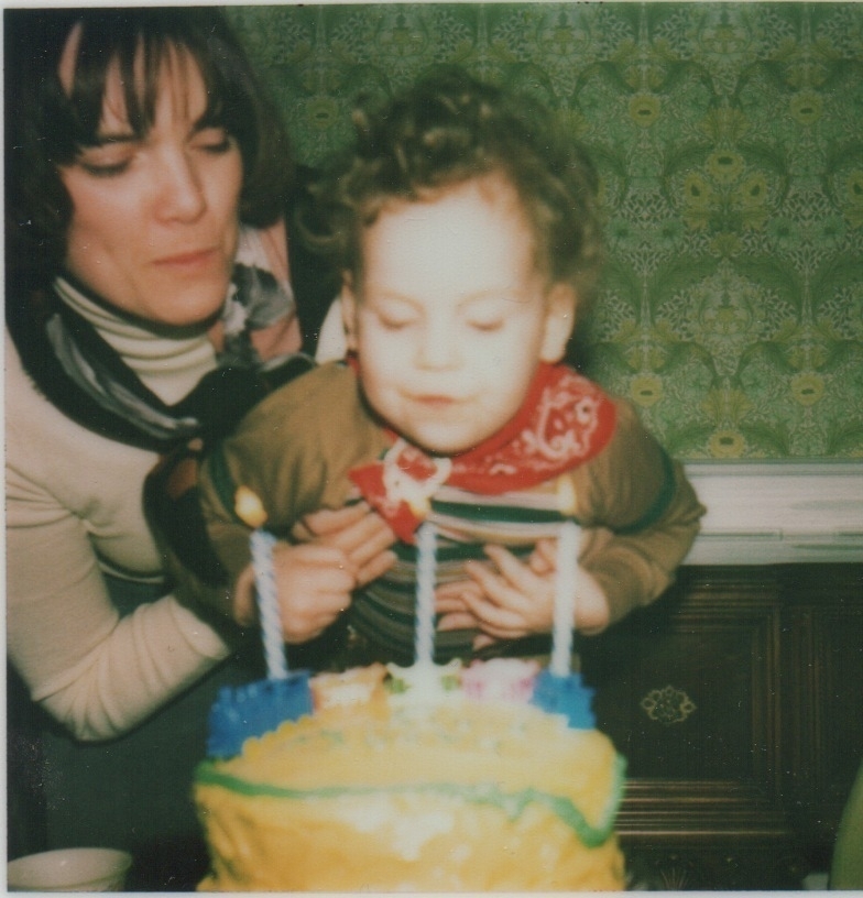 A child blows birthday candles on a cake, with a woman assisting, in a room with patterned wallpaper.