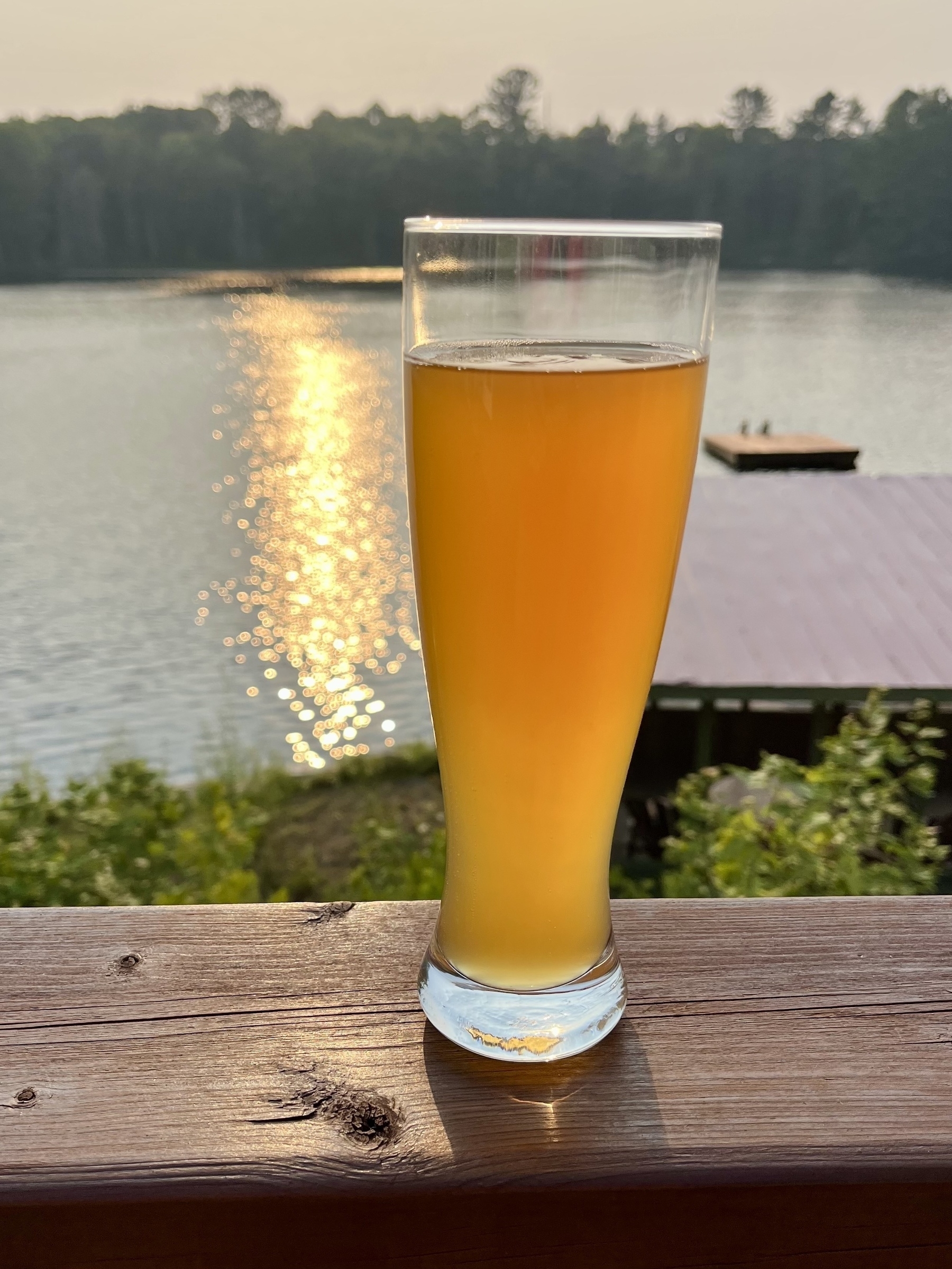 A tall glass of beer is placed on a wooden railing overlooking a lake with a sunset reflecting on the water.