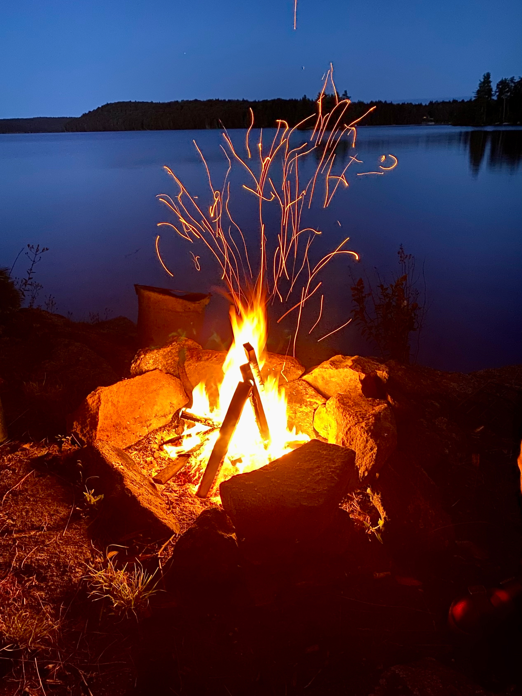 A campfire emits glowing sparks into the night sky by a tranquil lakeside.