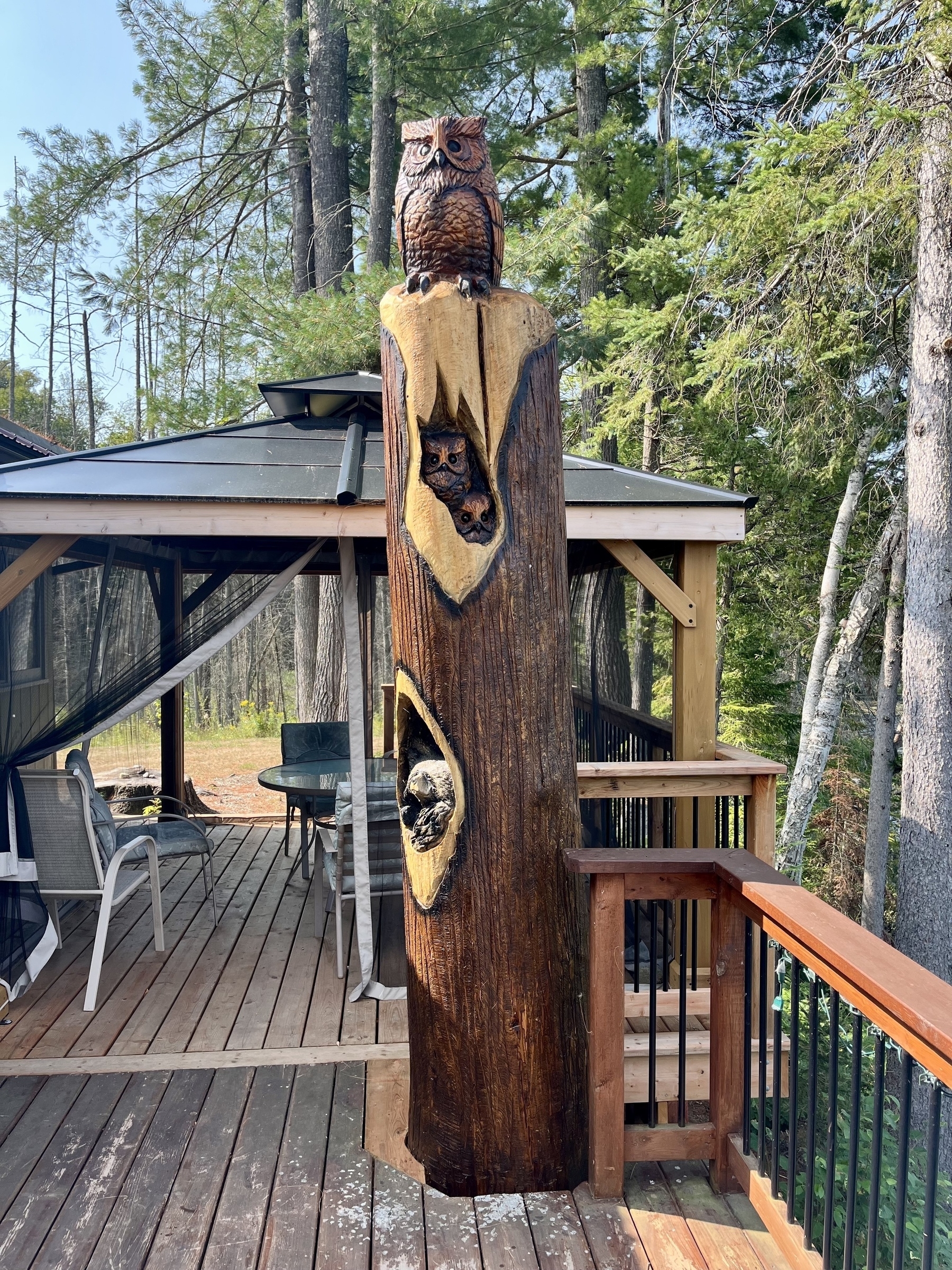 A wooden totem pole carved with animal figures, including an owl on top, stands on a deck next to an outdoor structure surrounded by trees.