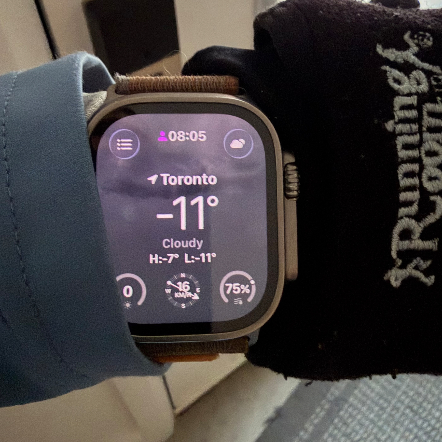 A person wearing a blue jacket and gloves is checking a smartwatch that displays the weather in Toronto as -11°C and cloudy.