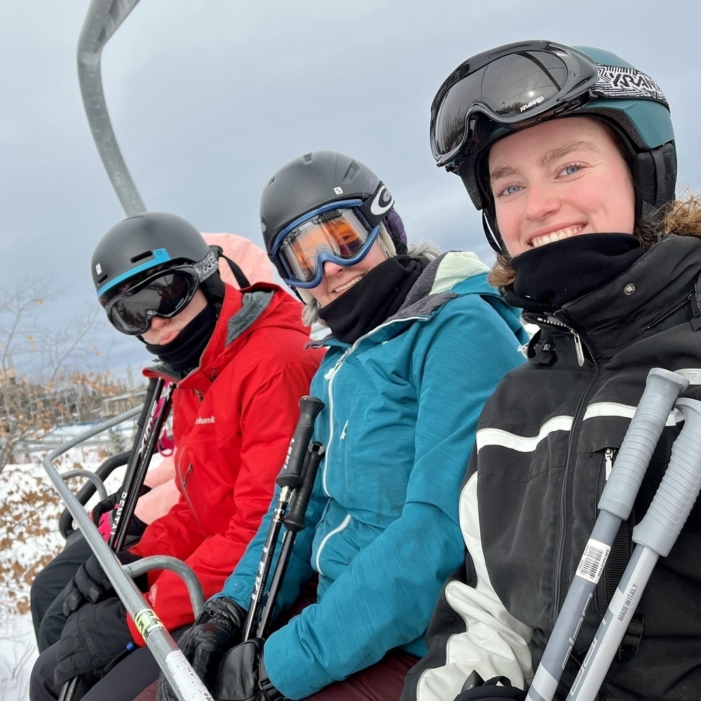 Three people are sitting on a ski lift, dressed in winter clothing and ski gear, smiling at the camera.