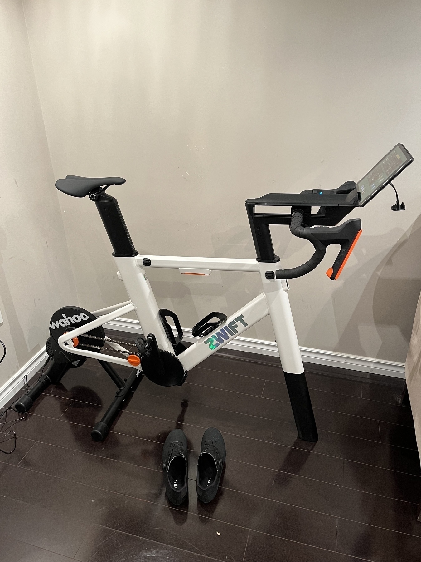 A stationary exercise bike with a "Zwift" logo stands in a room, accompanied by a pair of black shoes on the floor.