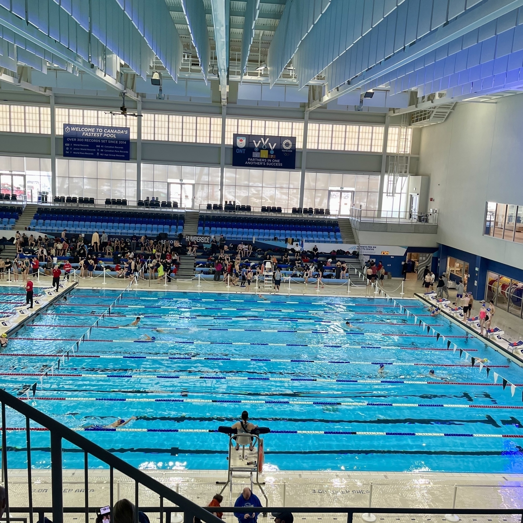 A busy indoor swimming pool hosts a swim meet, with swimmers in action and spectators in the stands.