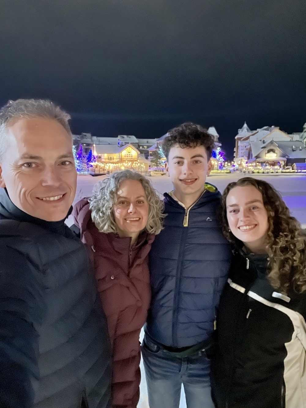 A family of four is smiling together at a snowy, festive outdoor location with lit-up buildings in the background.