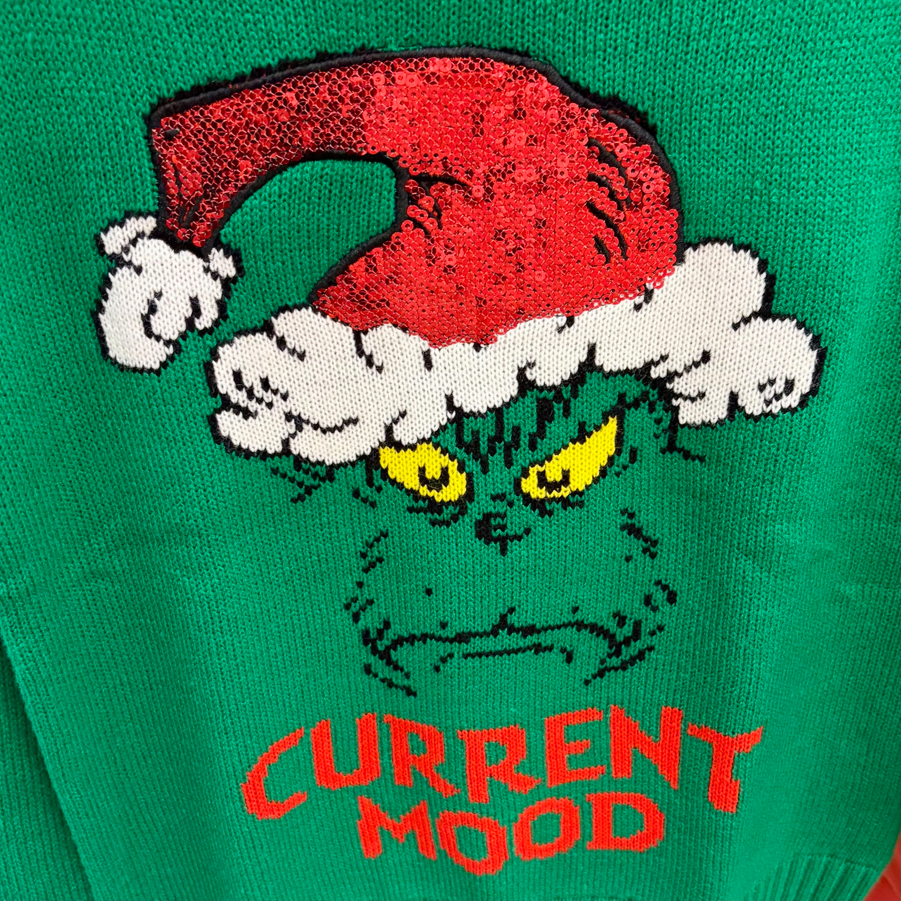 A green sweater features a grumpy face wearing a Santa hat with the words CURRENT MOOD below it.