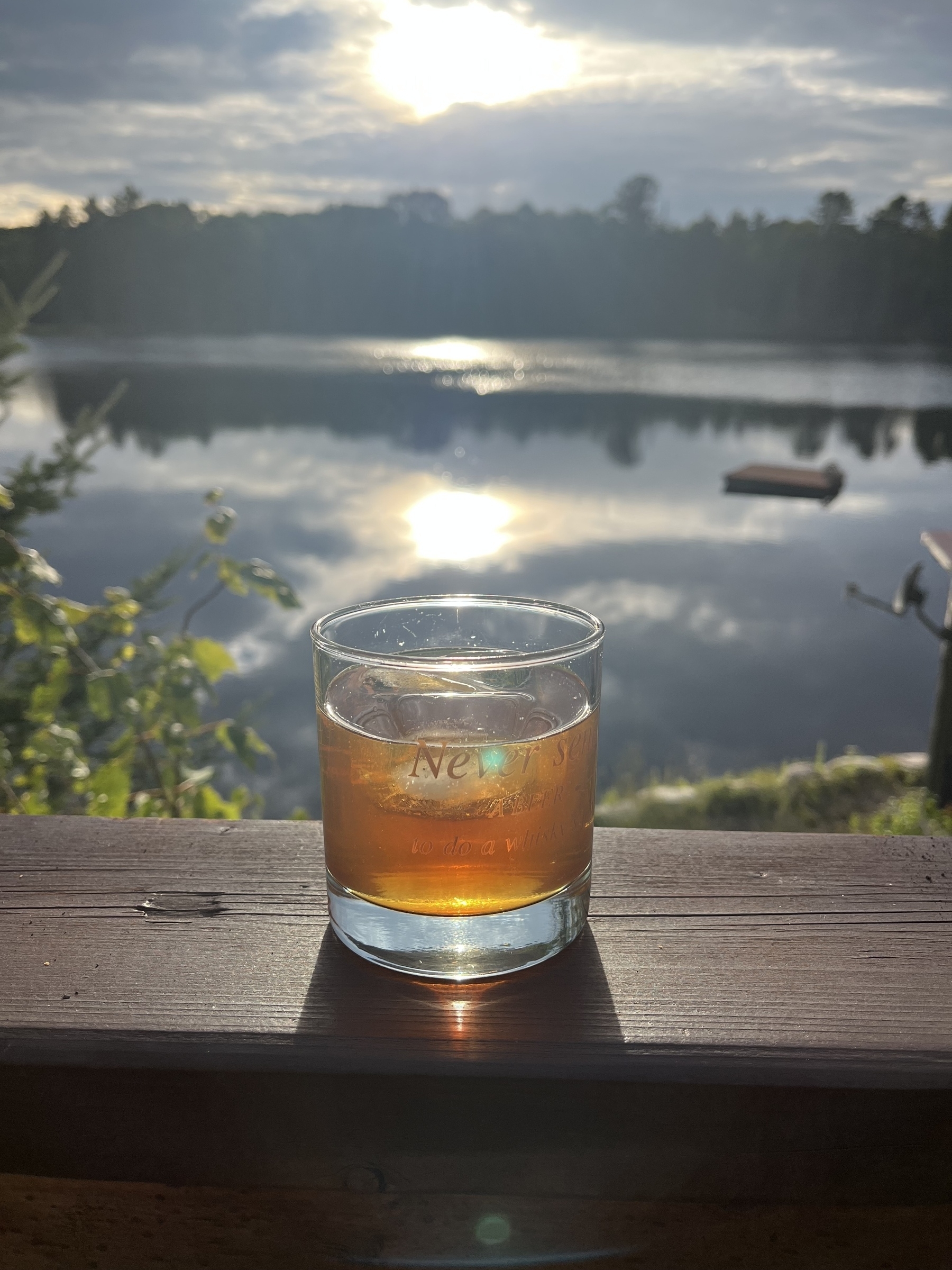 A glass filled with an amber liquid sits on a wooden railing overlooking a serene lake with the sun reflecting on the water.