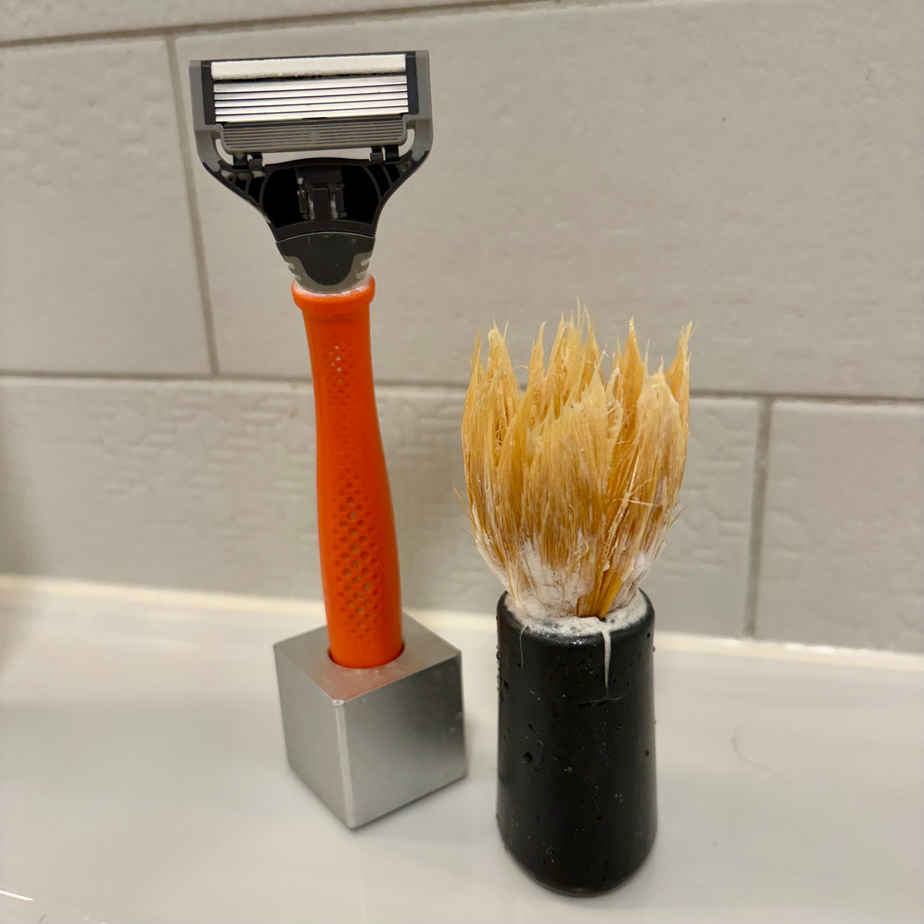 A razor with an orange handle is next to a shaving brush with a black base on a bathroom counter.