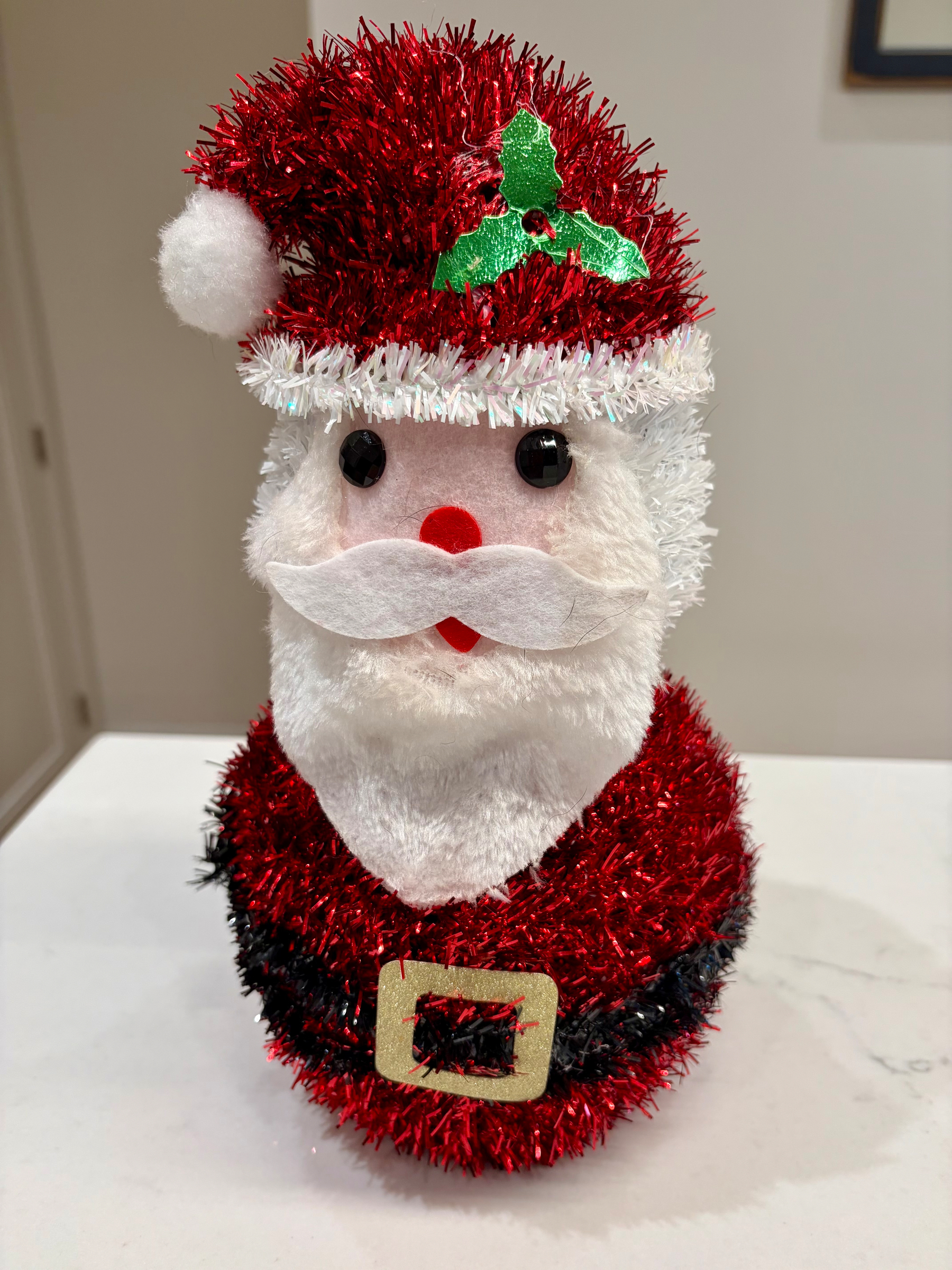 A festive Santa Claus decoration features a red and white tinsel outfit with a green holly accent on the hat.
