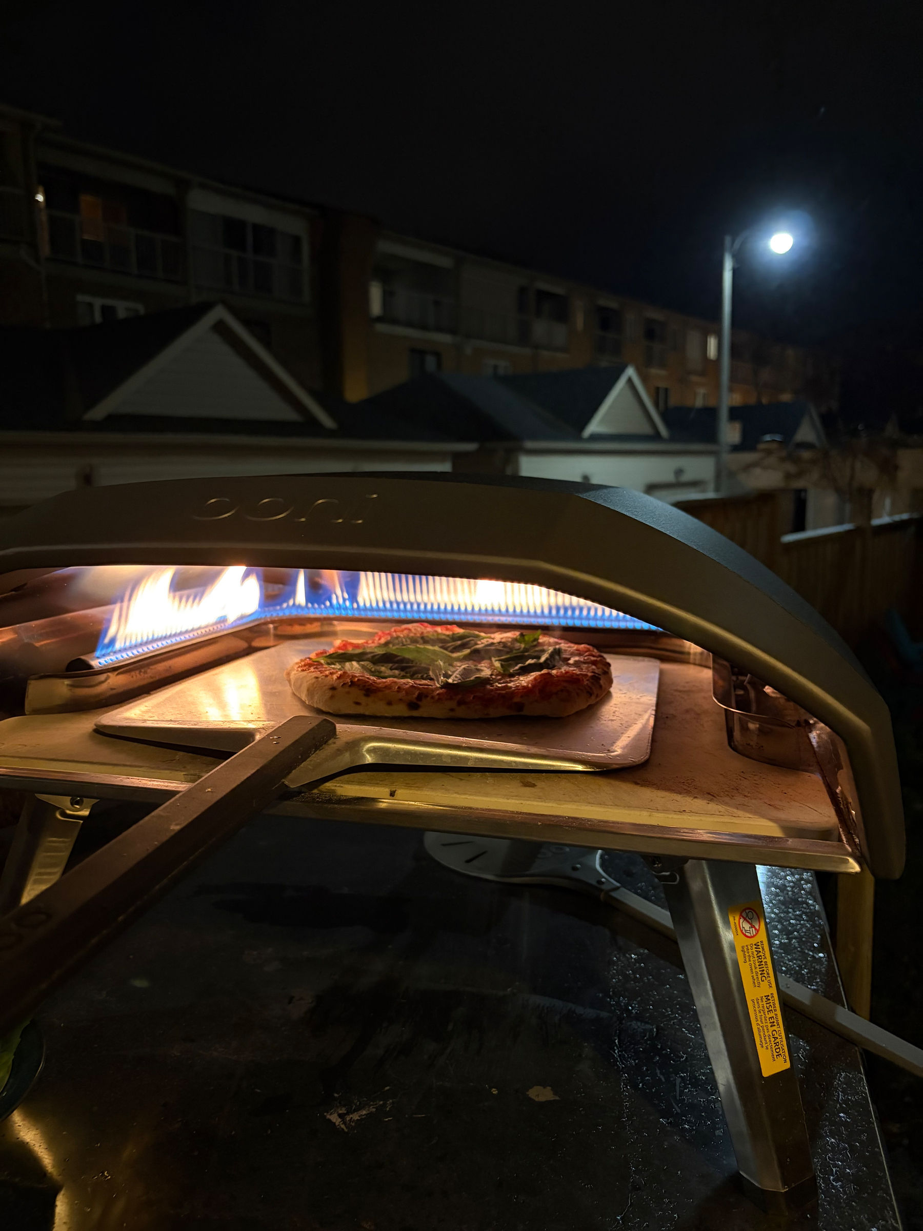 A freshly baked pizza is being cooked in an outdoor pizza oven, illuminated by its internal flame, against a nighttime residential backdrop.