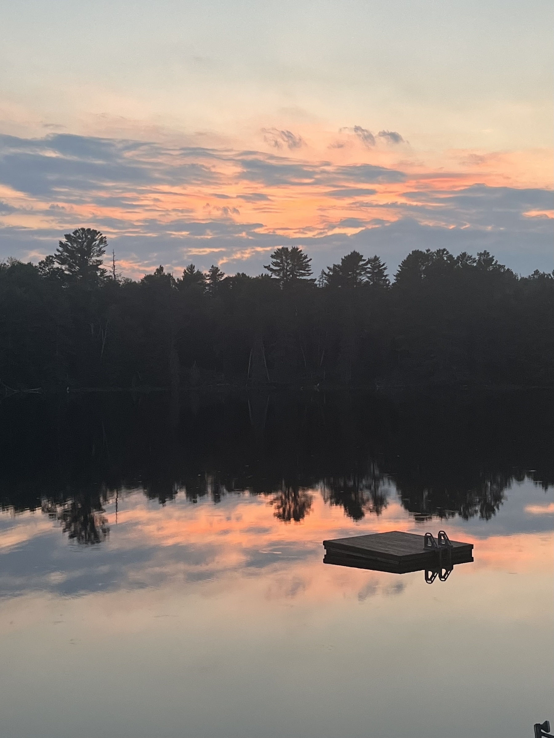 A serene lake reflects the pastel hues of a sunset sky, with a small floating dock visible in the water.