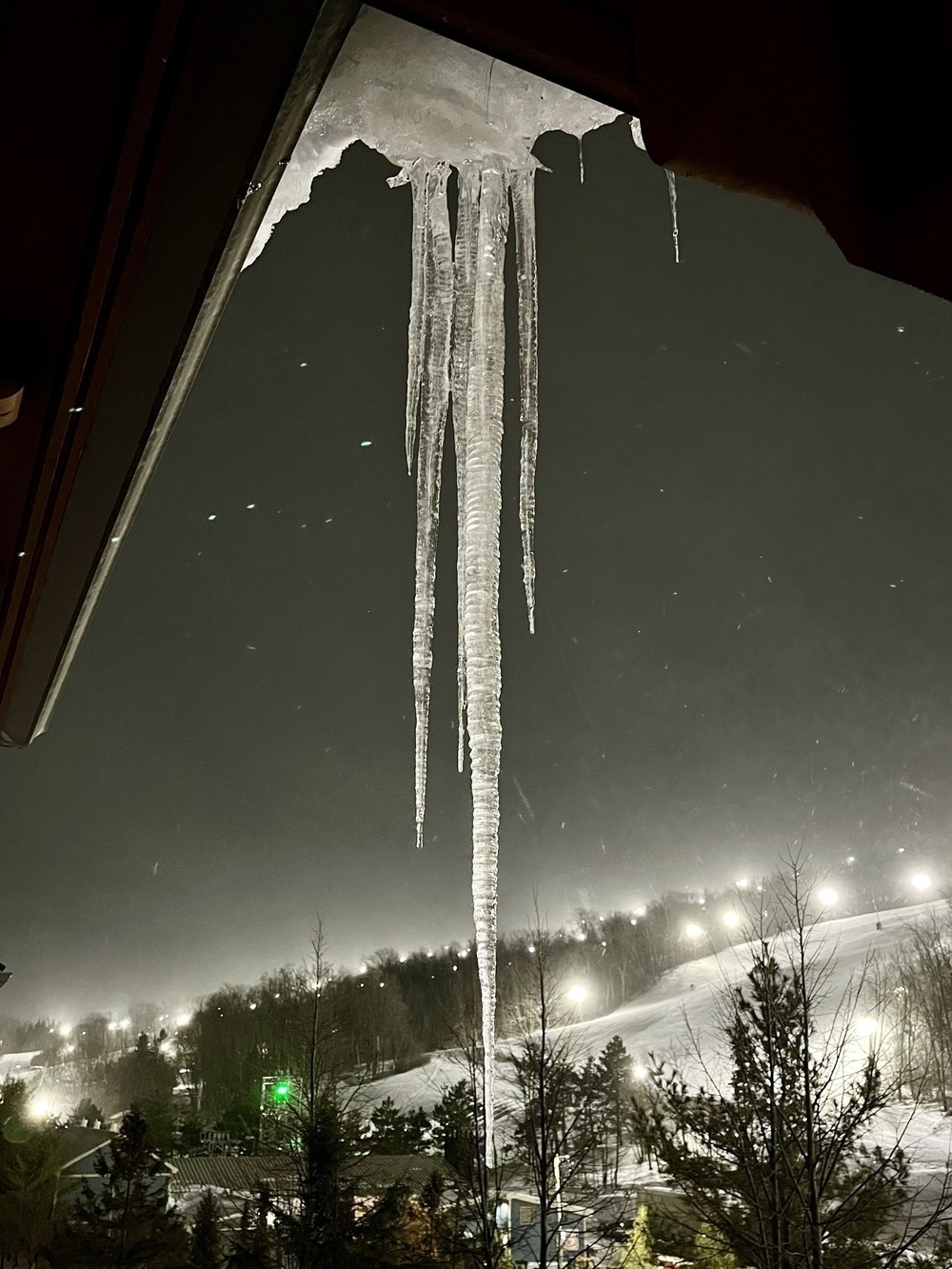 Long icicles hang from a rooftop with snow-covered trees and a softly lit ski slope in the background.