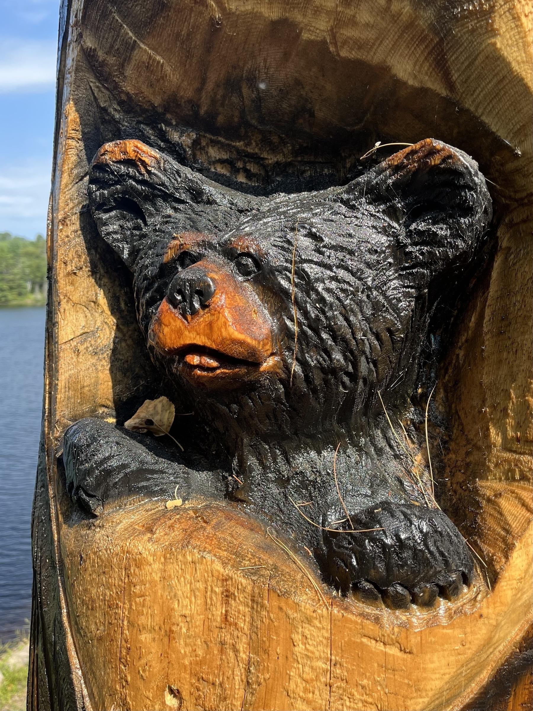 A wooden sculpture depicts the head of a bear, carved into a tree trunk, situated near a lake.