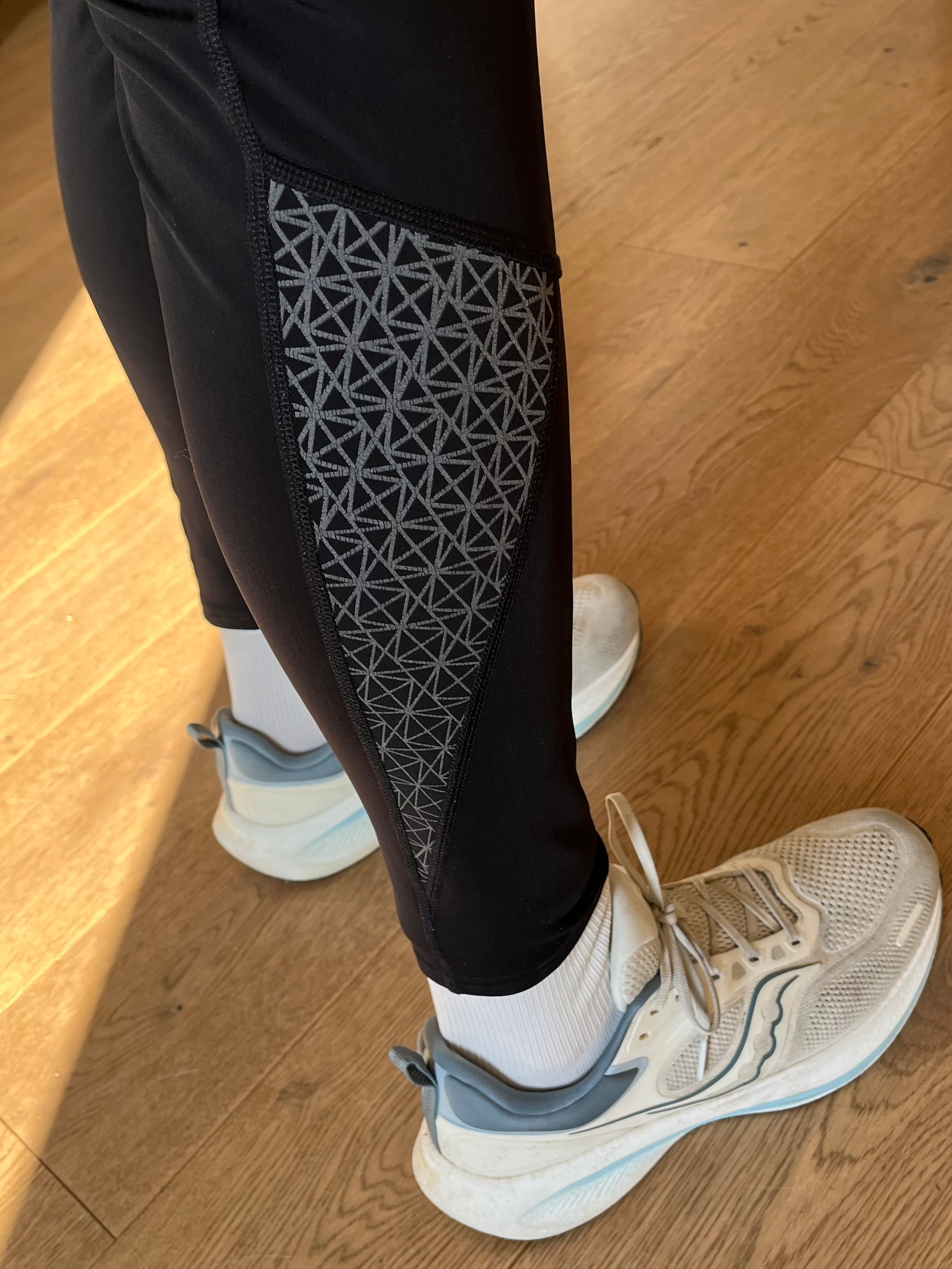 A person wearing black leggings with a geometric pattern on the sides is standing on a wooden floor, wearing white and gray athletic shoes and white socks.