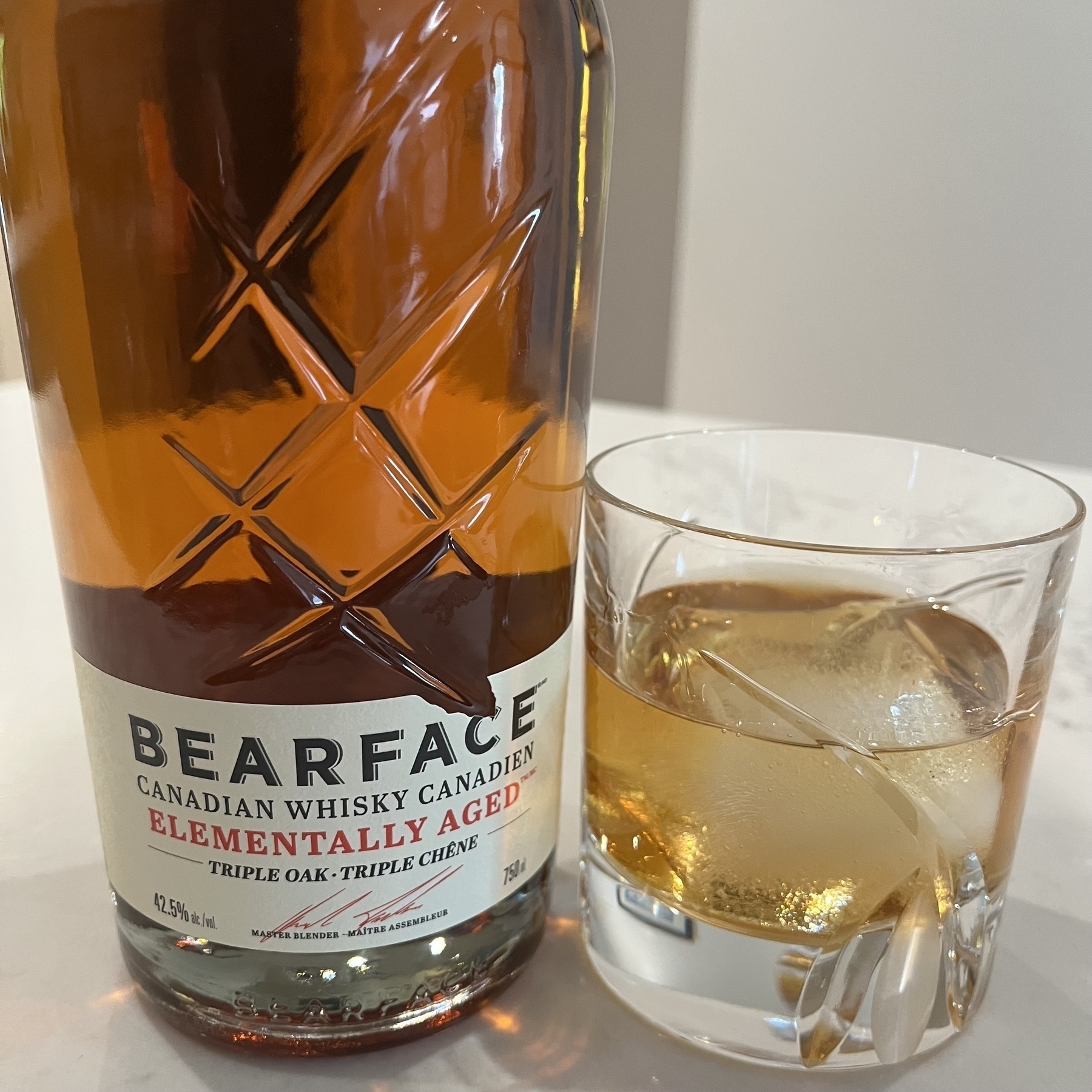 A bottle of Bearface Canadian Whisky stands next to a glass filled with whisky and a large ice cube.