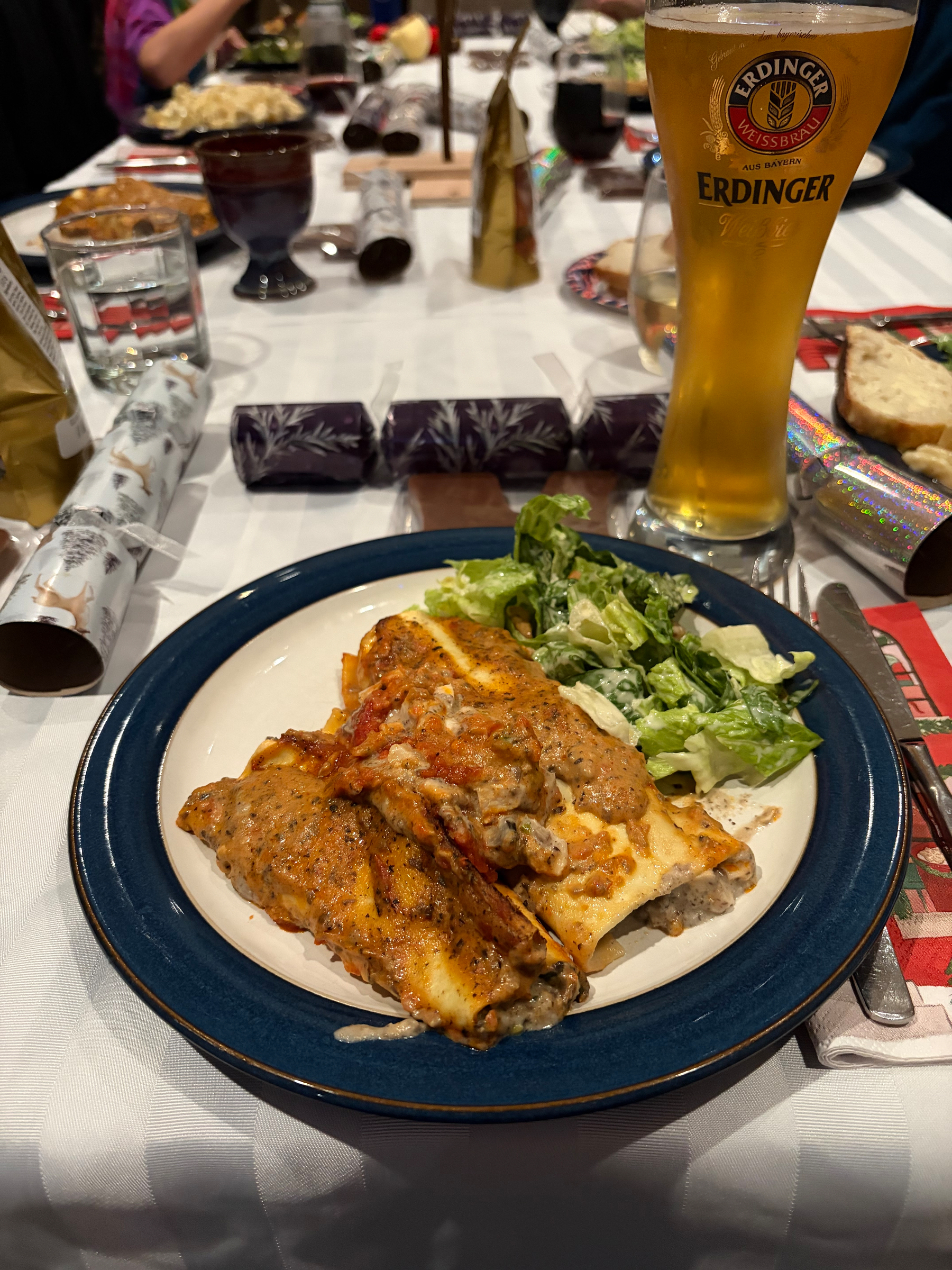 A plate of cannelloni with salad is served alongside a glass of Erdinger beer on a table set for a meal.