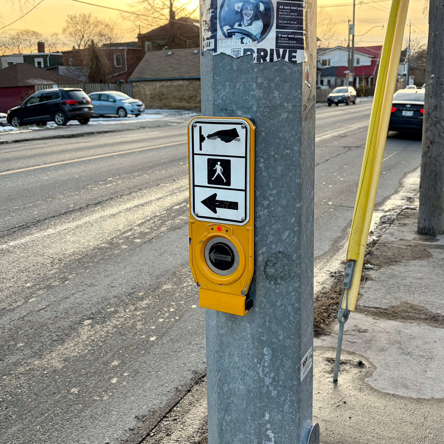 A pedestrian crossing button is attached to a pole beside a road with houses and cars in the background.