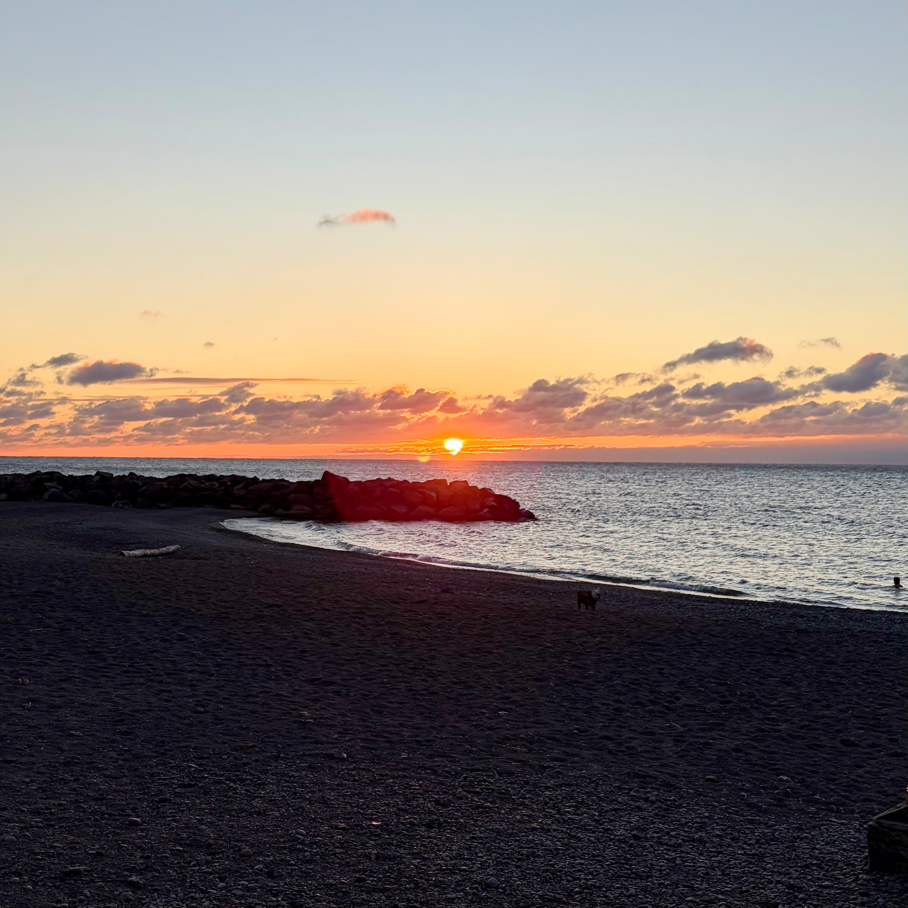 A serene sunset over the ocean with a rocky shoreline and a dark sandy beach.