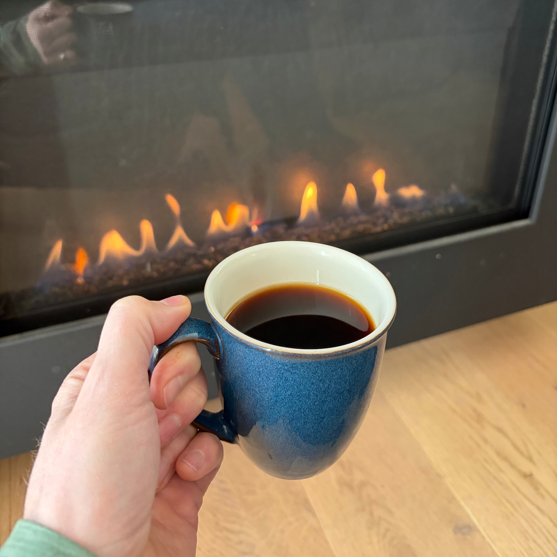 A hand holds a blue mug of coffee in front of a lit fireplace on a wooden floor.