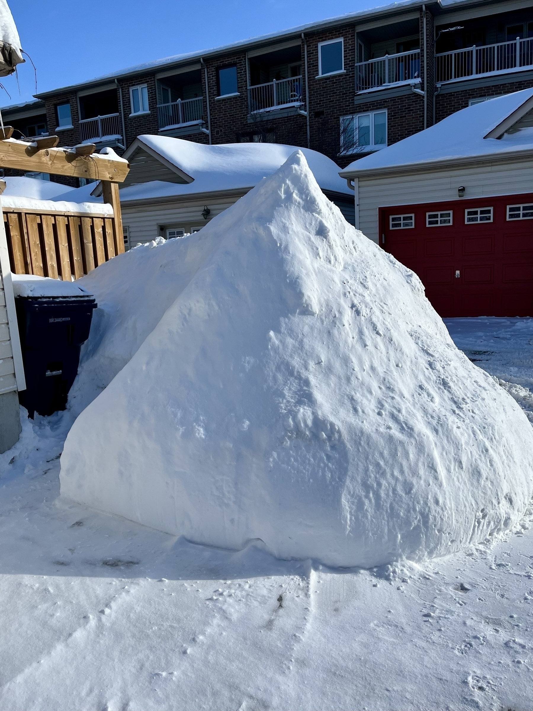 A large pile of snow is seen in front of houses with red garage doors in a residential area.