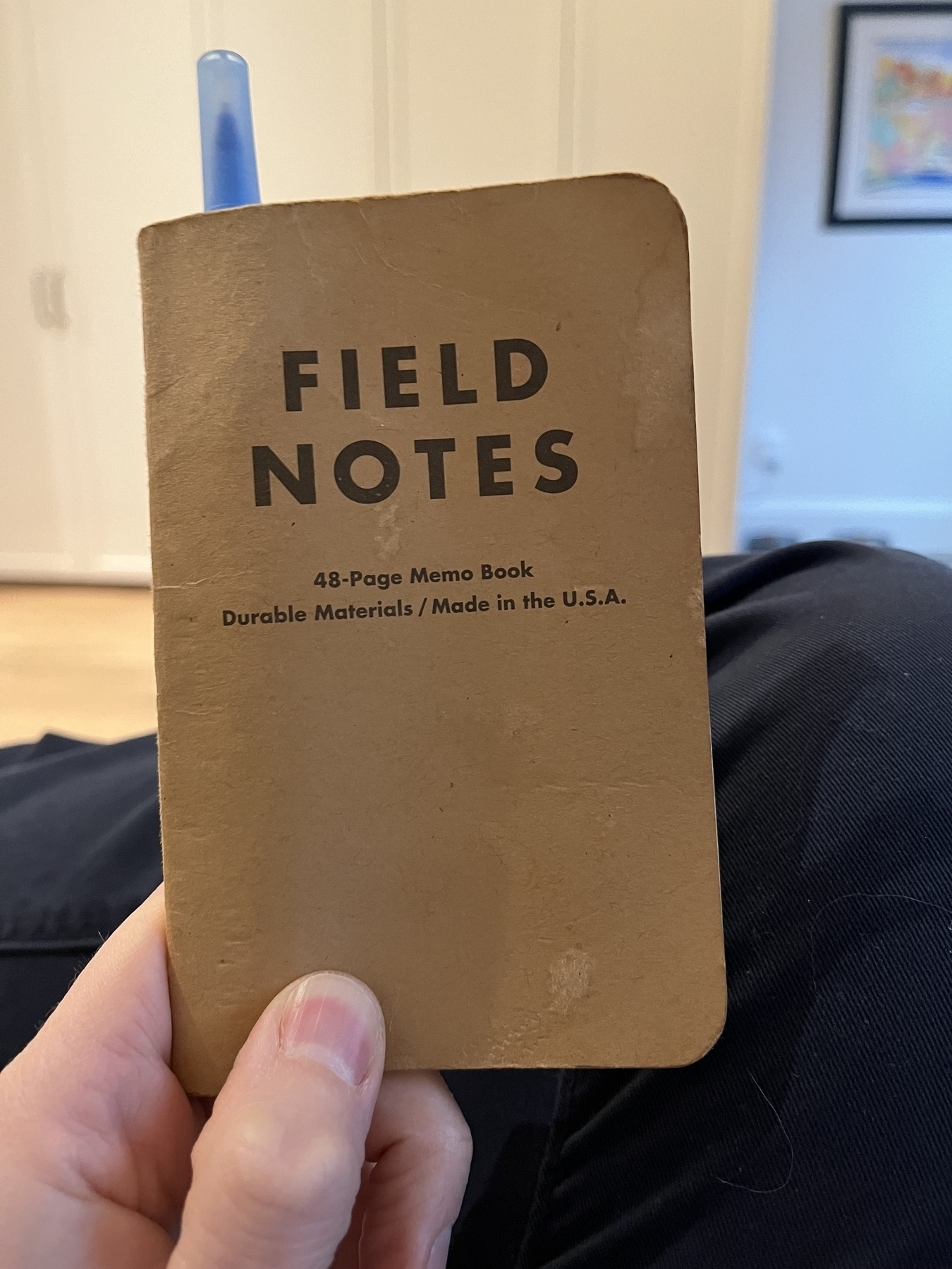 A hand holds a brown Field Notes memo book with a blue pen clipped to it.