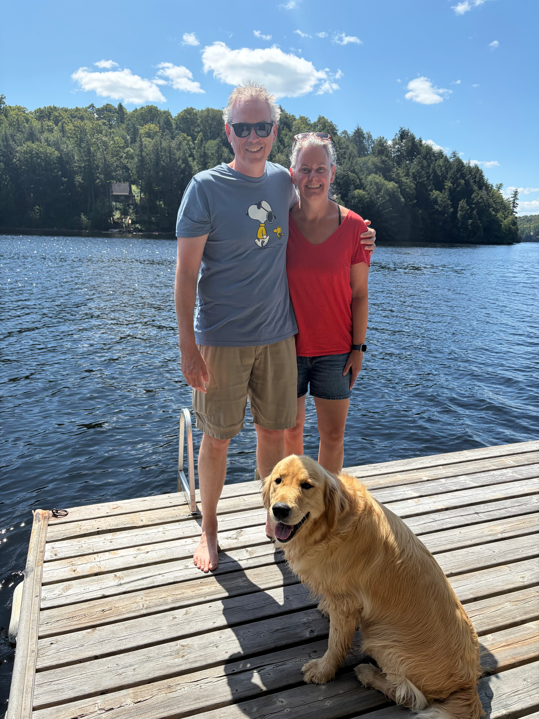 Auto-generated description: A smiling couple stands on a wooden dock by a lake with a golden retriever sitting in front of them on a sunny day.