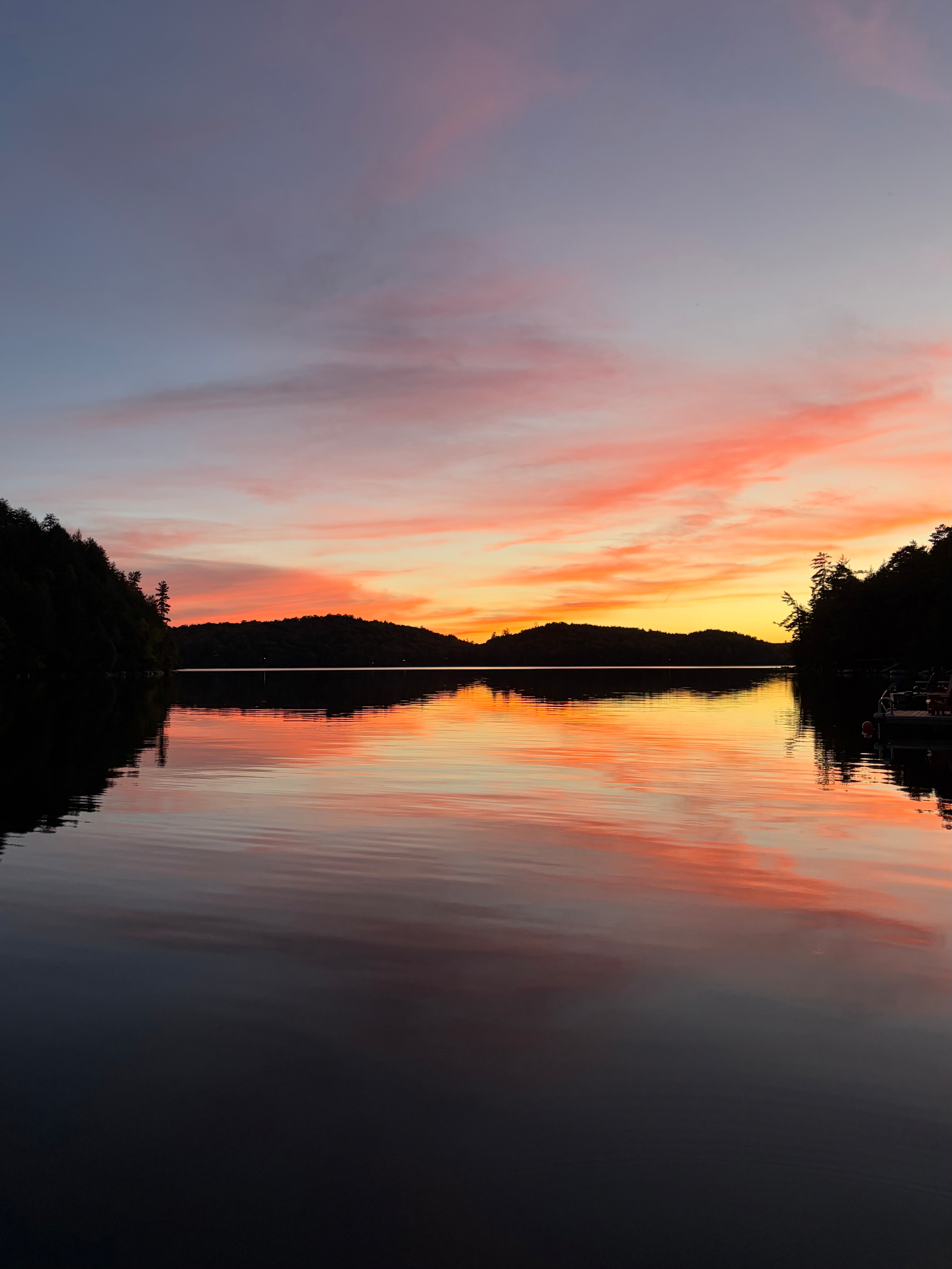 A tranquil lake reflects the vibrant colors of a sunset sky, bordered by dark silhouettes of trees and hills.