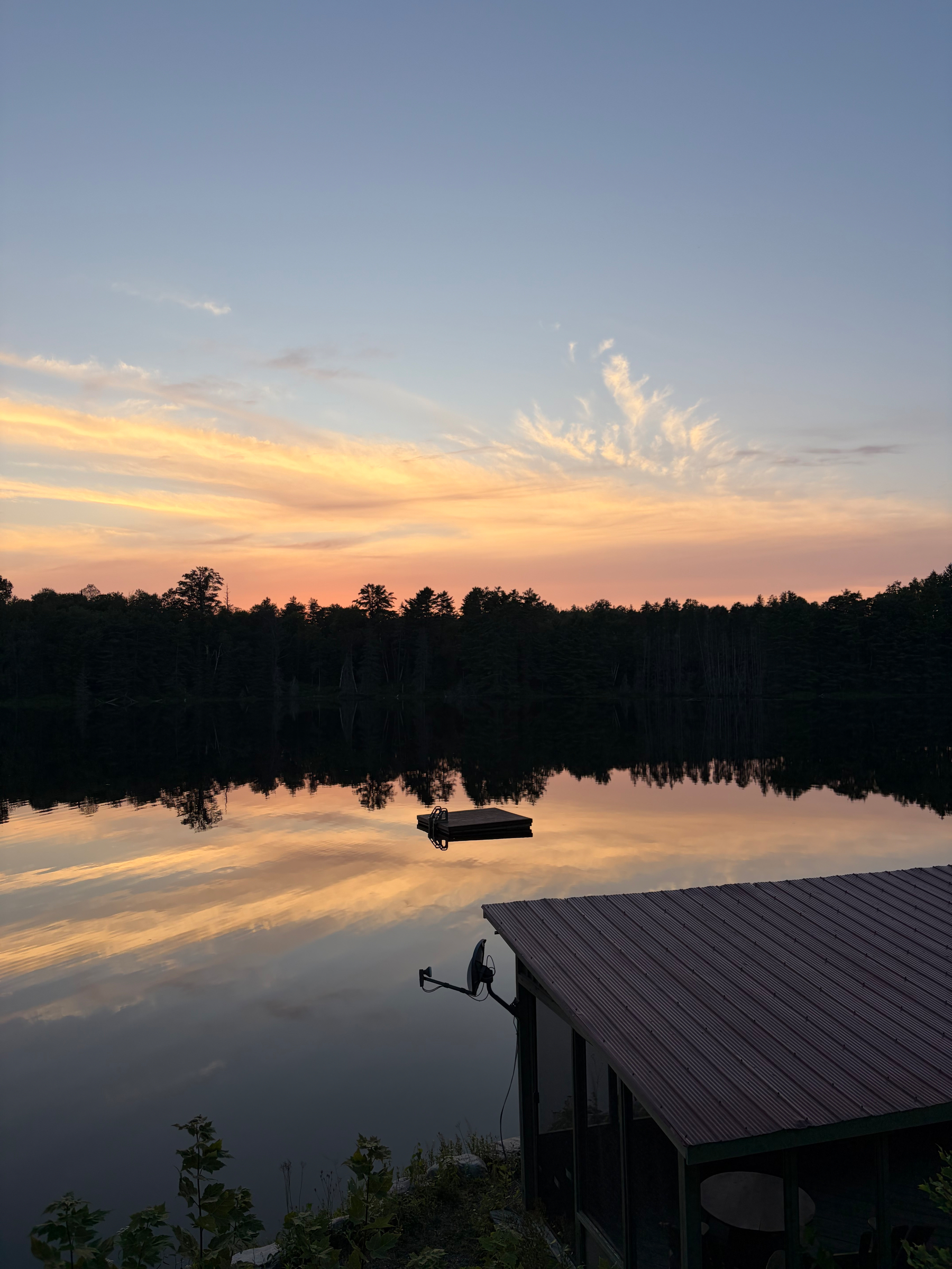 A calm lake at sunset reflects the sky with soft clouds, surrounded by trees, and features a small dock and a floating platform.