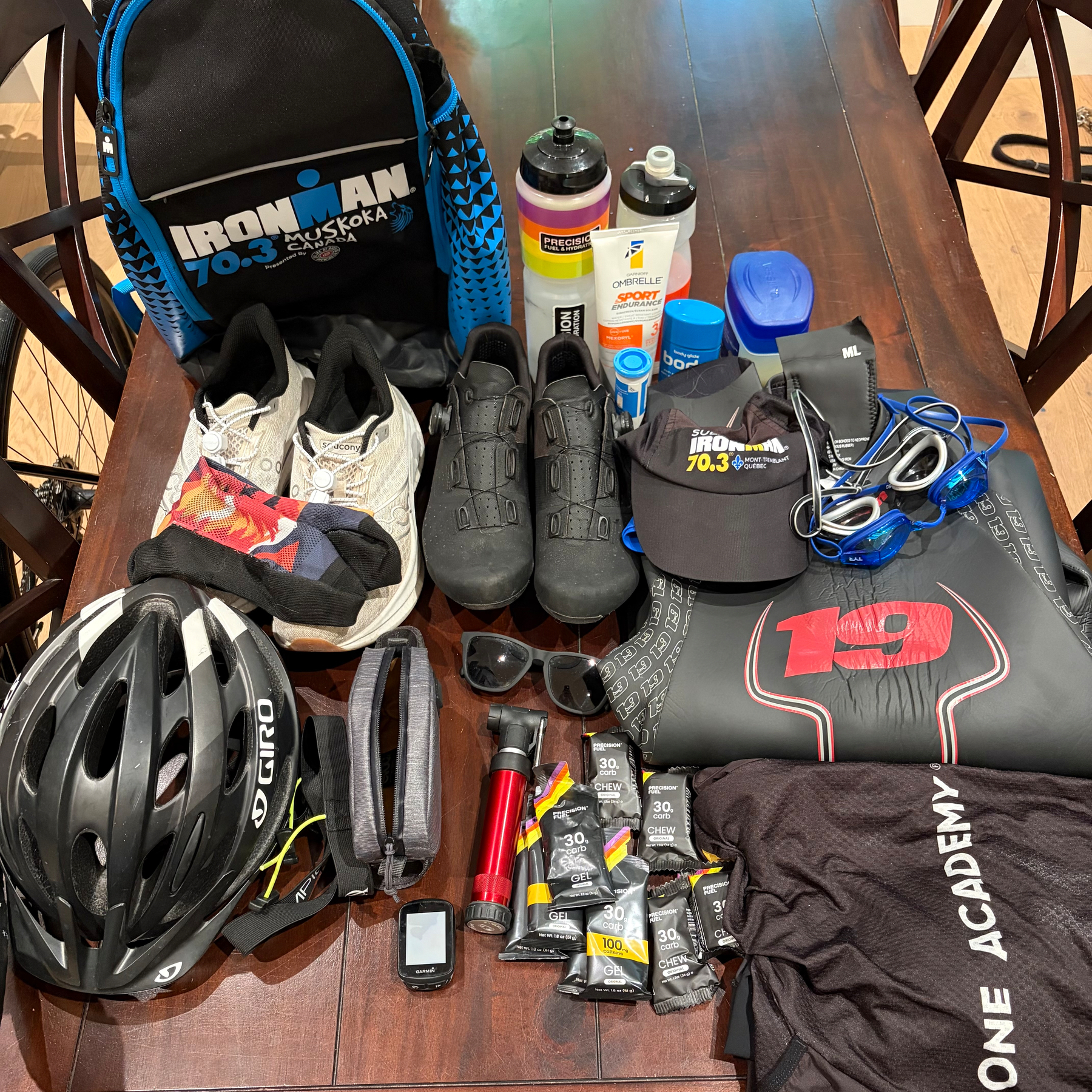 A collection of triathlon gear including a helmet, cycling shoes, running shoes, water bottles, nutrition gels, sunglasses, a cycling computer, swim goggles, and a backpack is arranged on a wooden table.