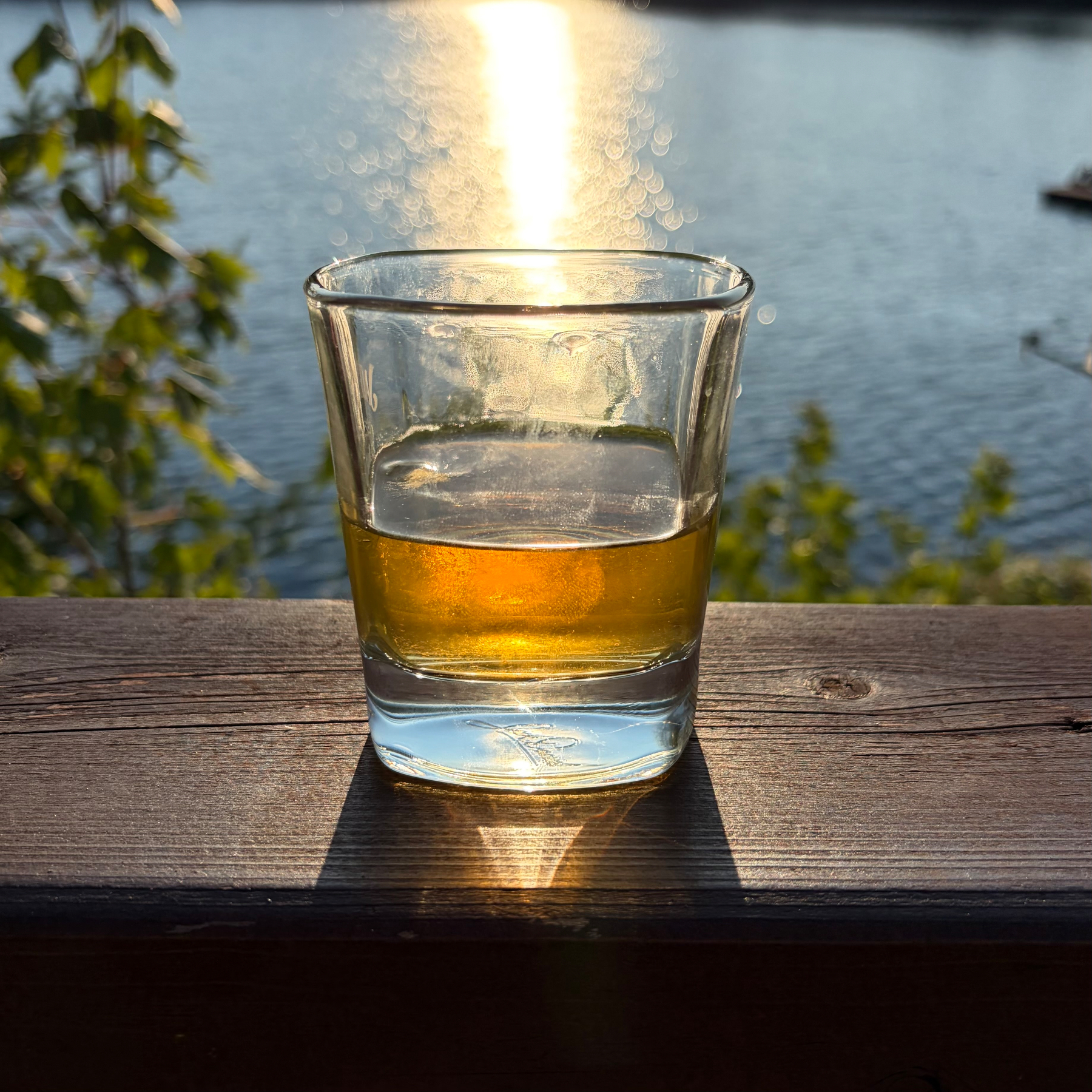 A glass with a golden liquid is placed on a wooden surface with sunlight reflecting off a body of water in the background.