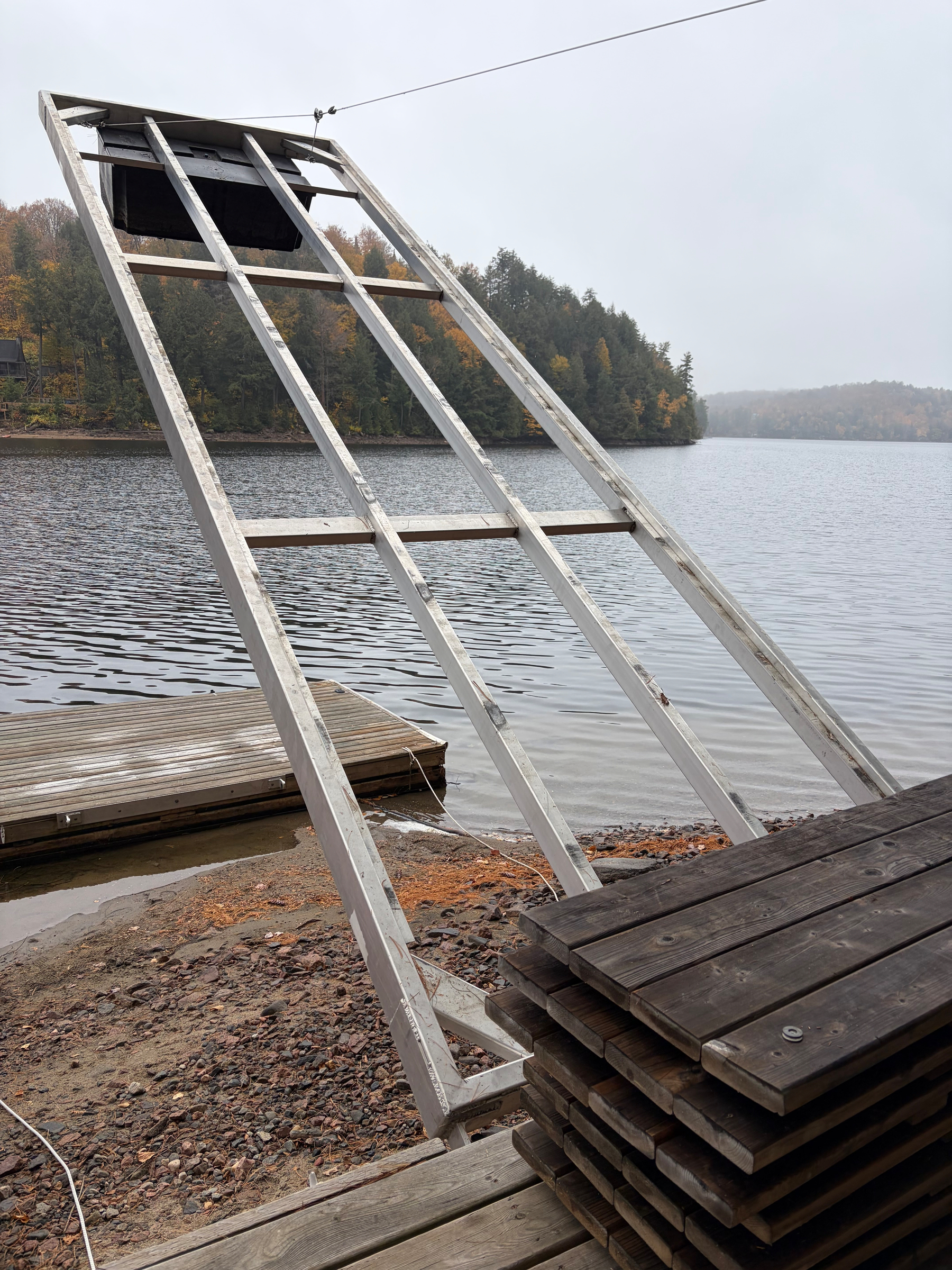 A large wooden ramp structure extends from a stack of wooden planks near a lakeshore, with trees and a small dock visible in the background.
