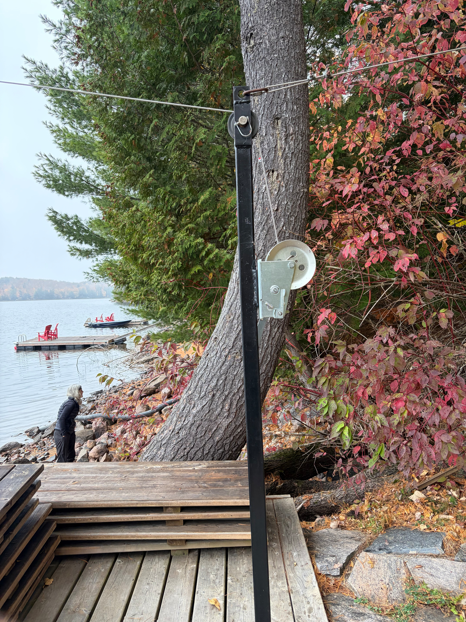 A metal cable system with a pulley is installed near a lakeside dock surrounded by autumn foliage and a person standing by the water.
