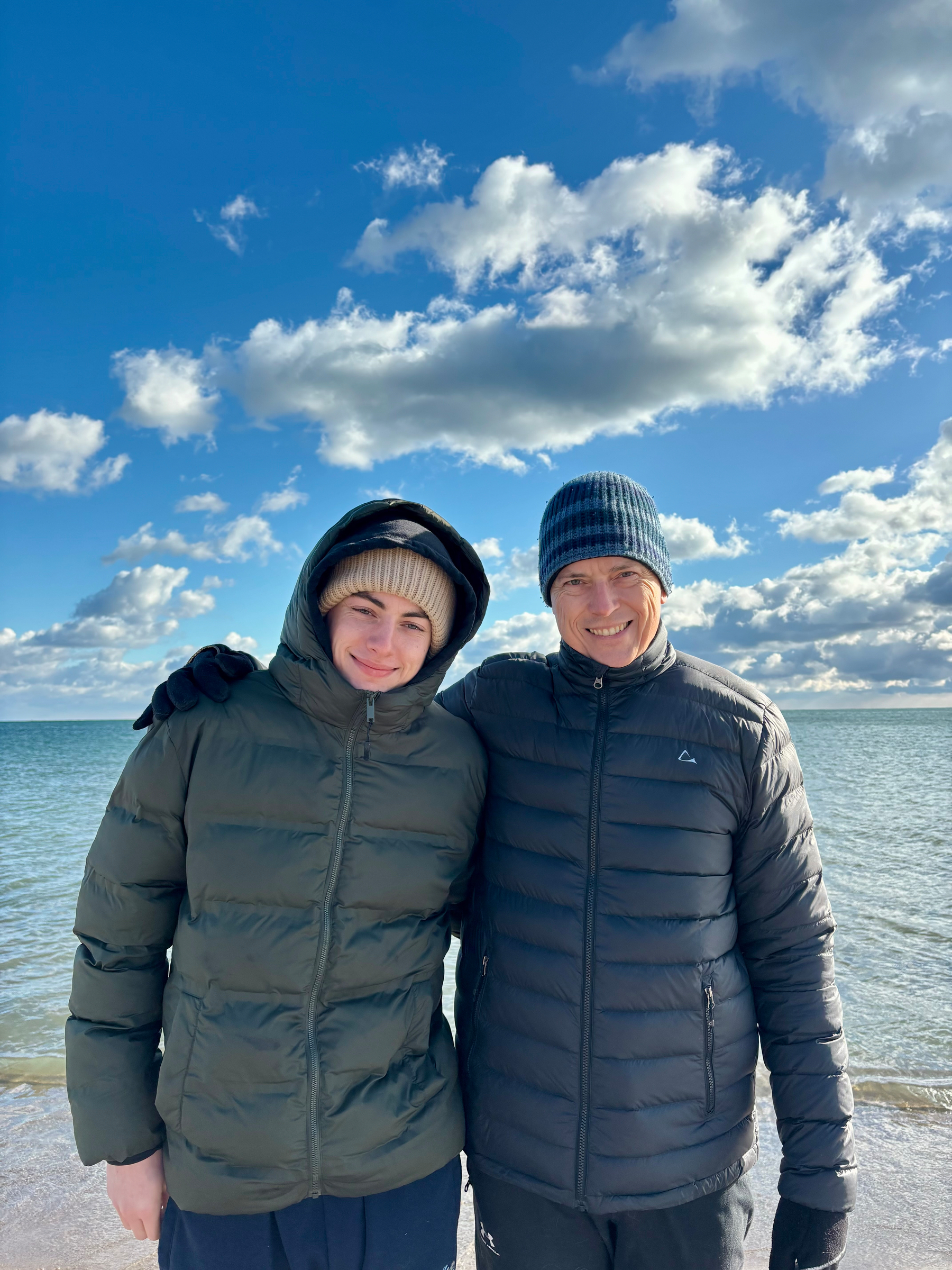 Two people wearing winter jackets and hats are standing on a beach with the lake and a cloudy sky in the background.