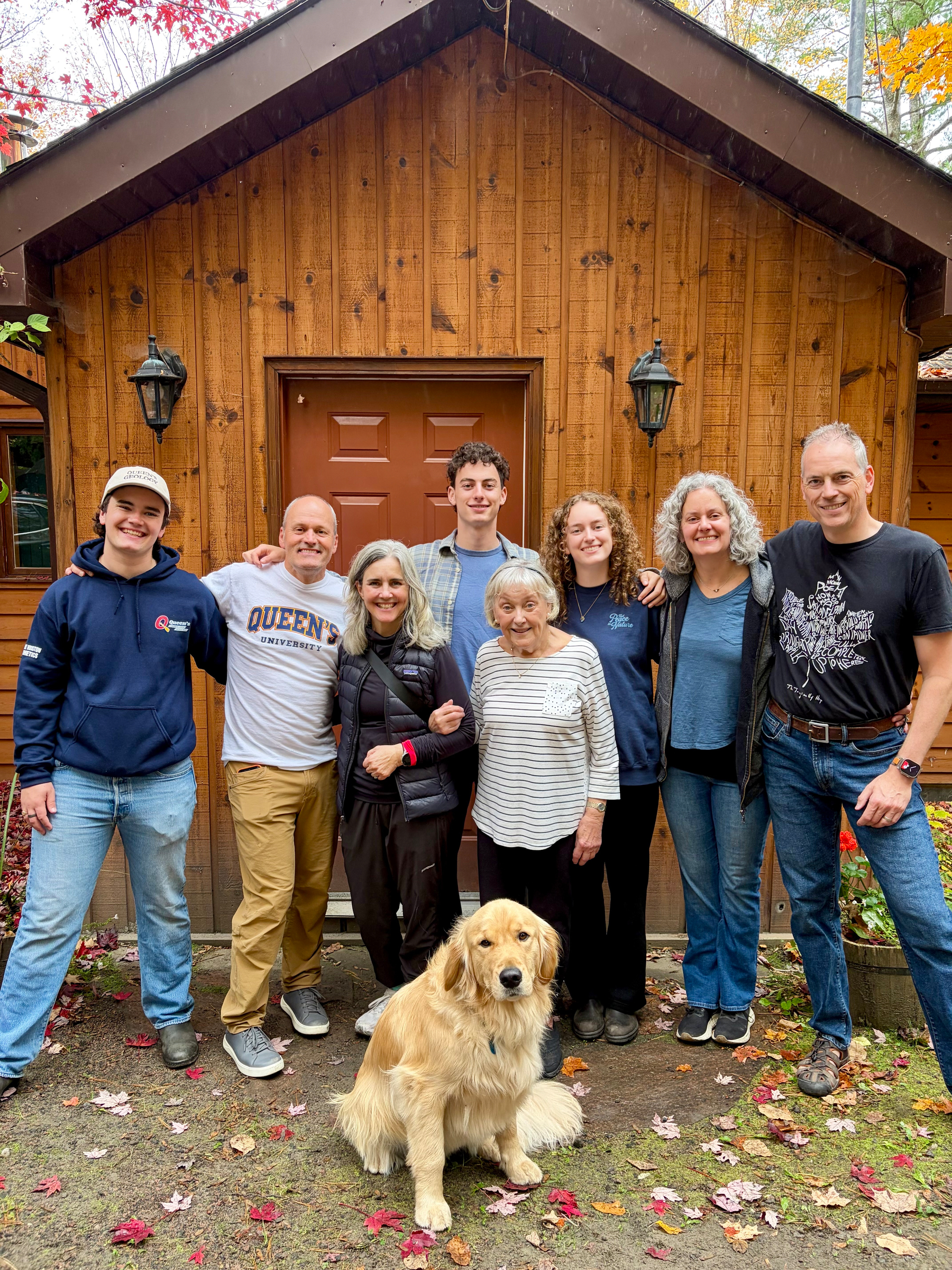 A group of seven people of different ages and a golden retriever are posing together in front of a wooden cabin door with autumn leaves scattered on the ground.