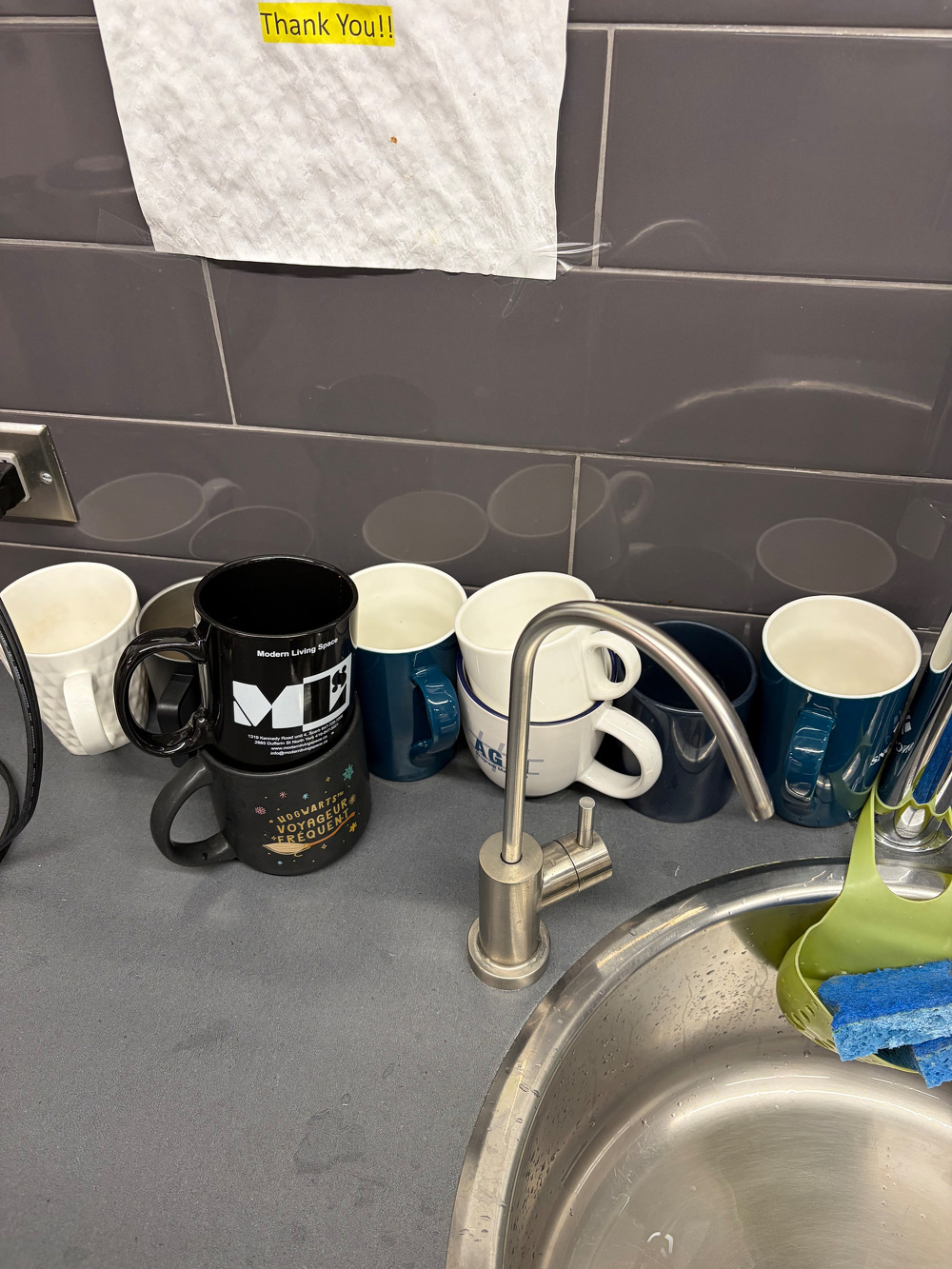 Several mugs are lined up on a kitchen counter near a sink with a spout and sponge.