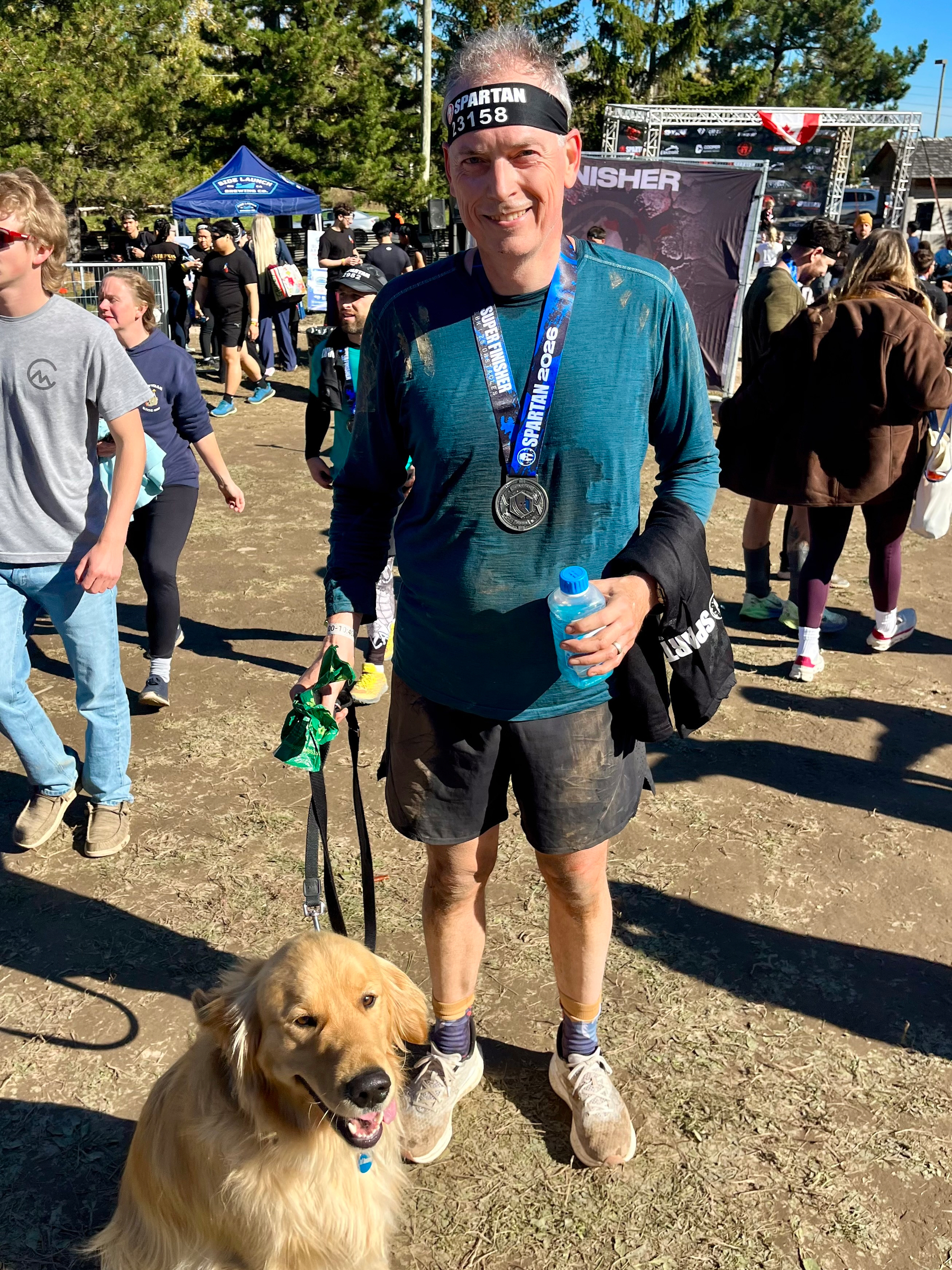 A man wearing a Spartan headband and medal is standing outdoors on a dirt ground with a golden retriever, holding a bottle of water, while people and event tents are visible in the background.