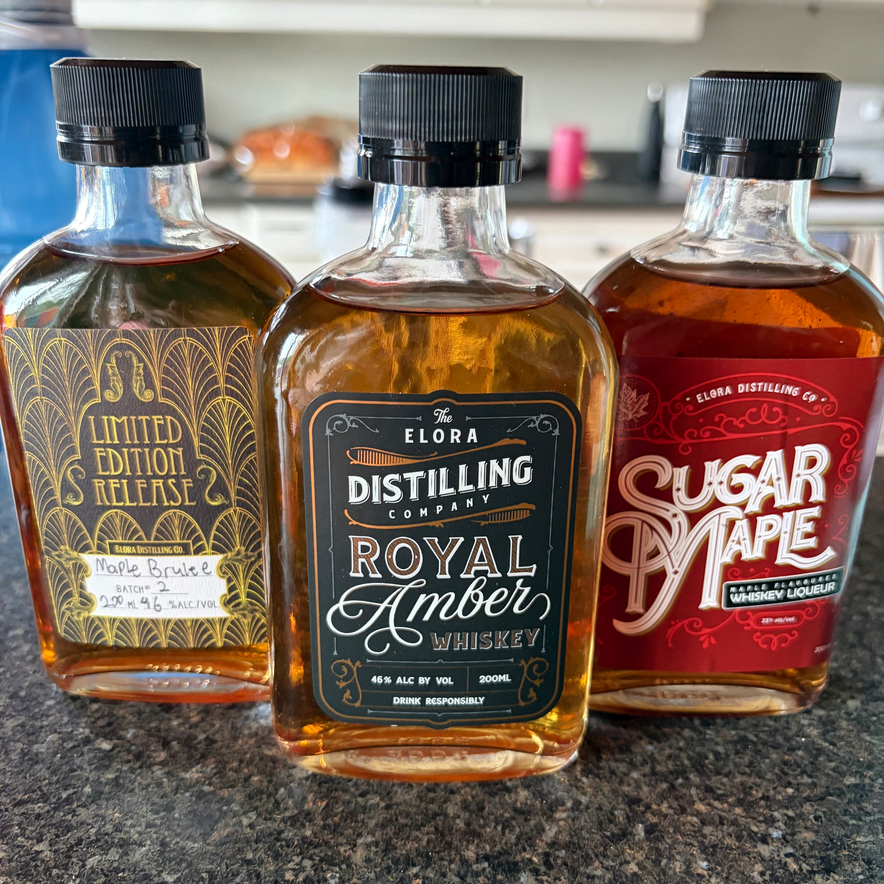 Three different bottles of whiskey liqueur from Elora Distilling Company, labeled as Limited Edition Release Maple Brulee, Royal Amber, and Sugar Maple, are displayed on a kitchen counter.