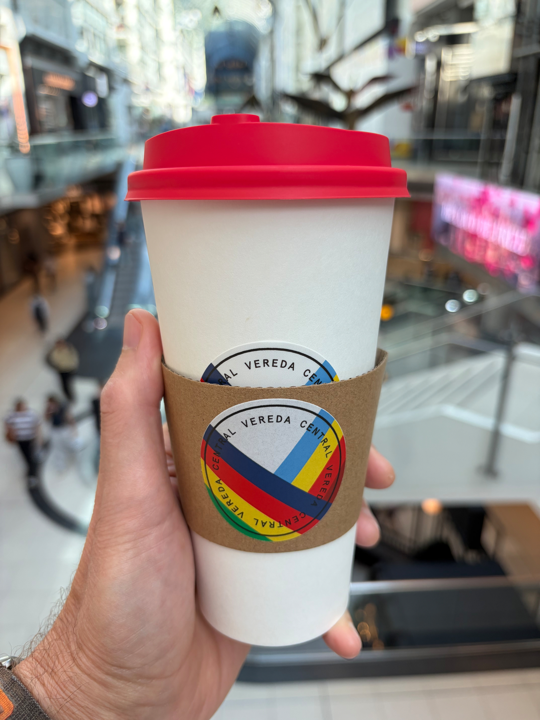 A hand is holding a white disposable coffee cup with a red lid and a cardboard sleeve that features a colorful circular logo with the text Vereda Central, inside a bright, modern shopping mall.