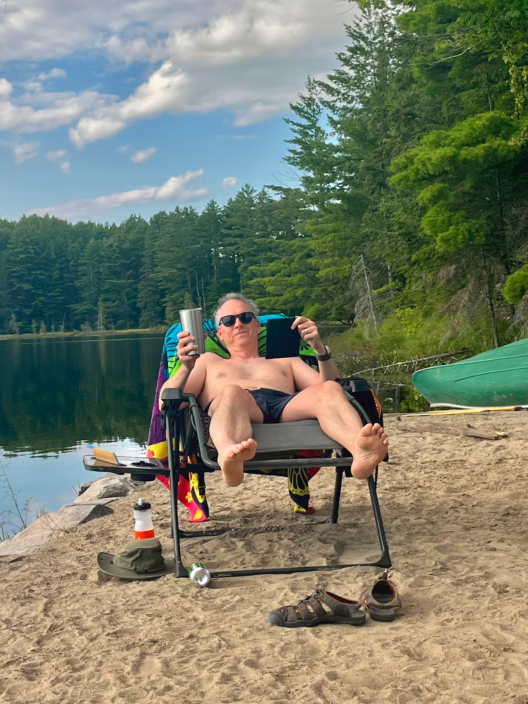 A man is relaxing shirtless on a beach chair by a lake, wearing sunglasses and holding a cup and a tablet, surrounded by trees and outdoor gear.