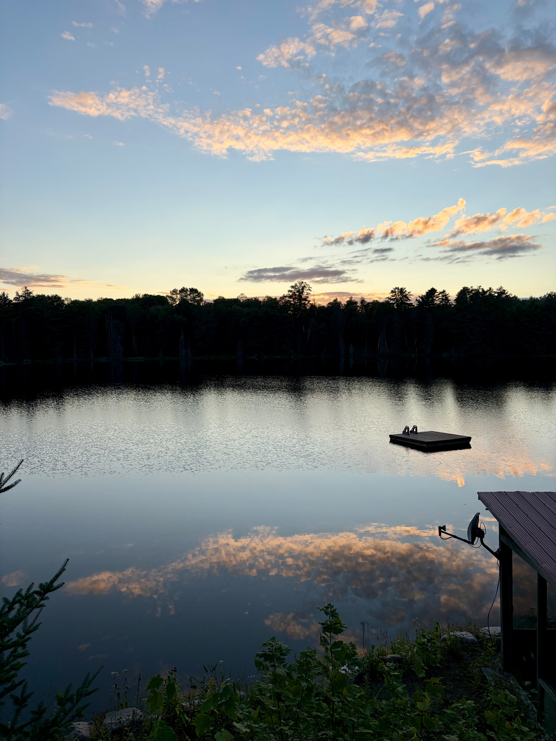 A serene lake reflects the cloudy sky at sunset, with a floating dock in the middle and a small pier on the right side.