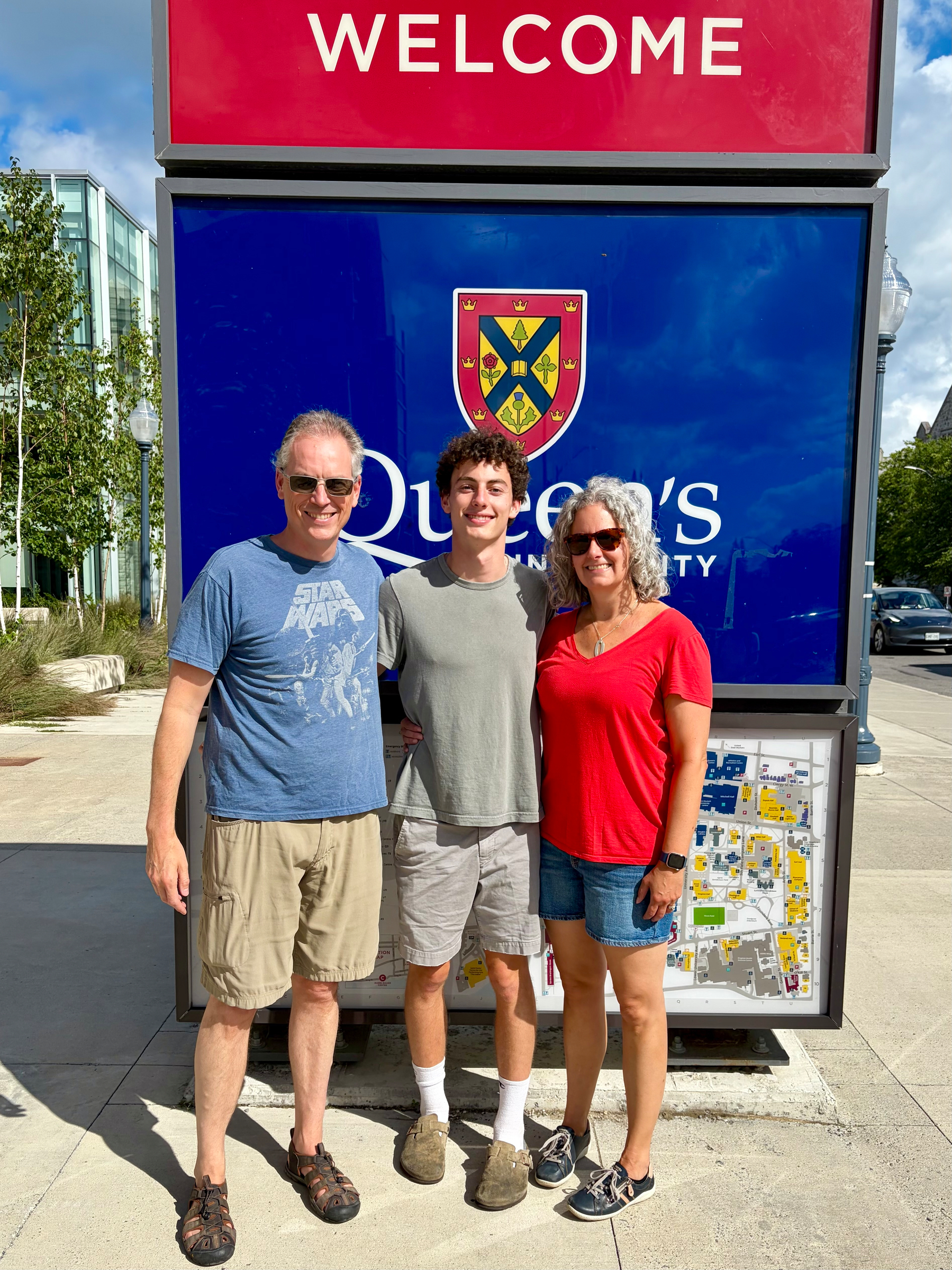 A group of three people stands smiling together in front of a Queen's University welcome sign on a sunny day.