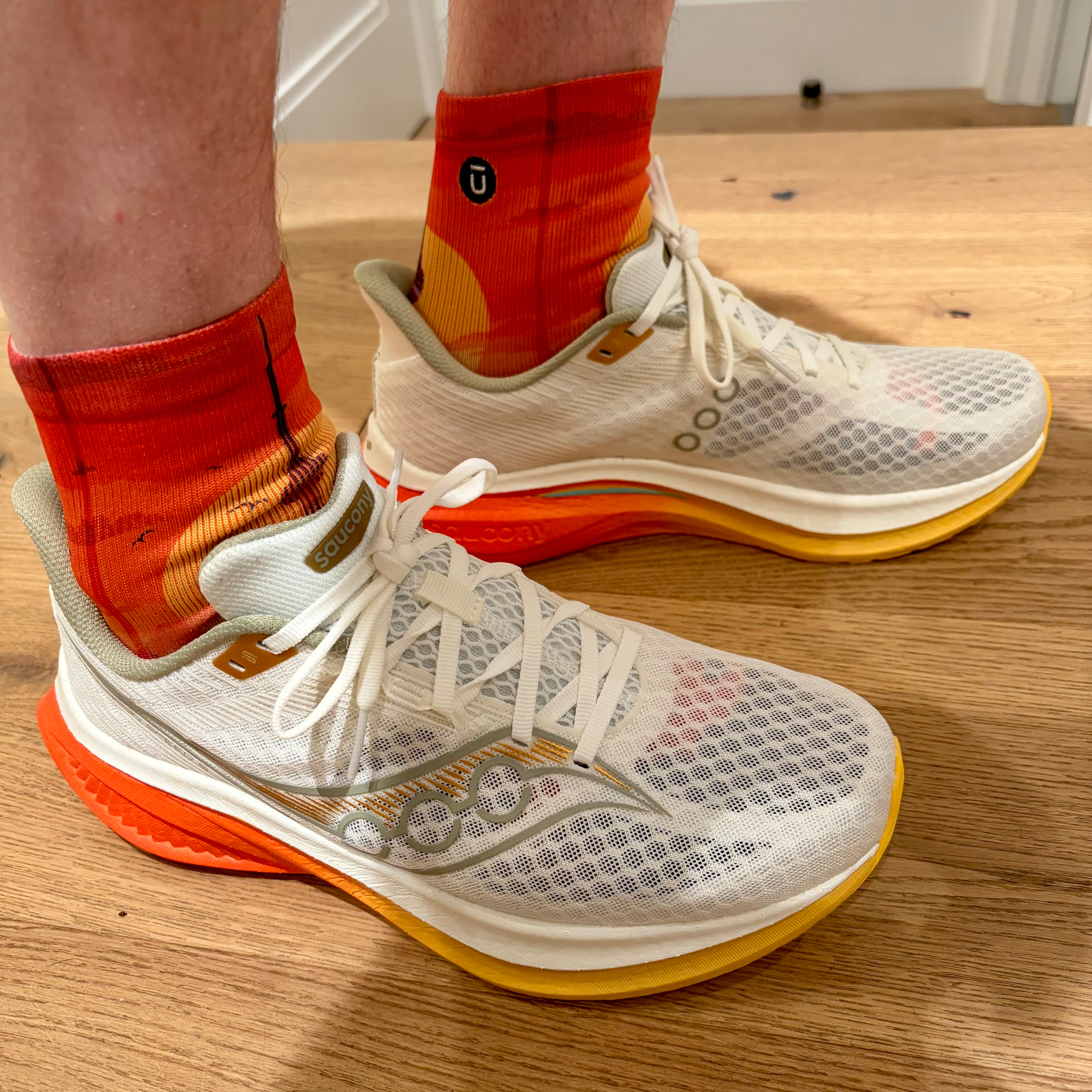 A pair of white and orange athletic shoes are being worn with red and orange socks standing on a wooden floor.