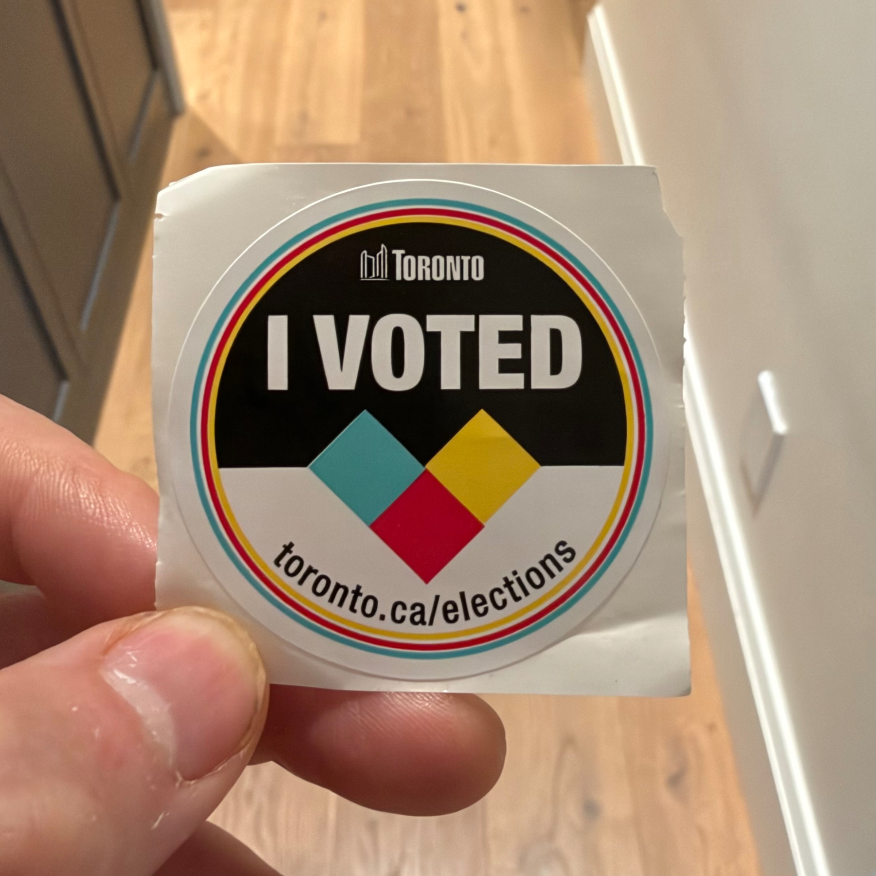 A hand is holding a circular sticker that says I VOTED with toronto.ca/elections printed beneath it.