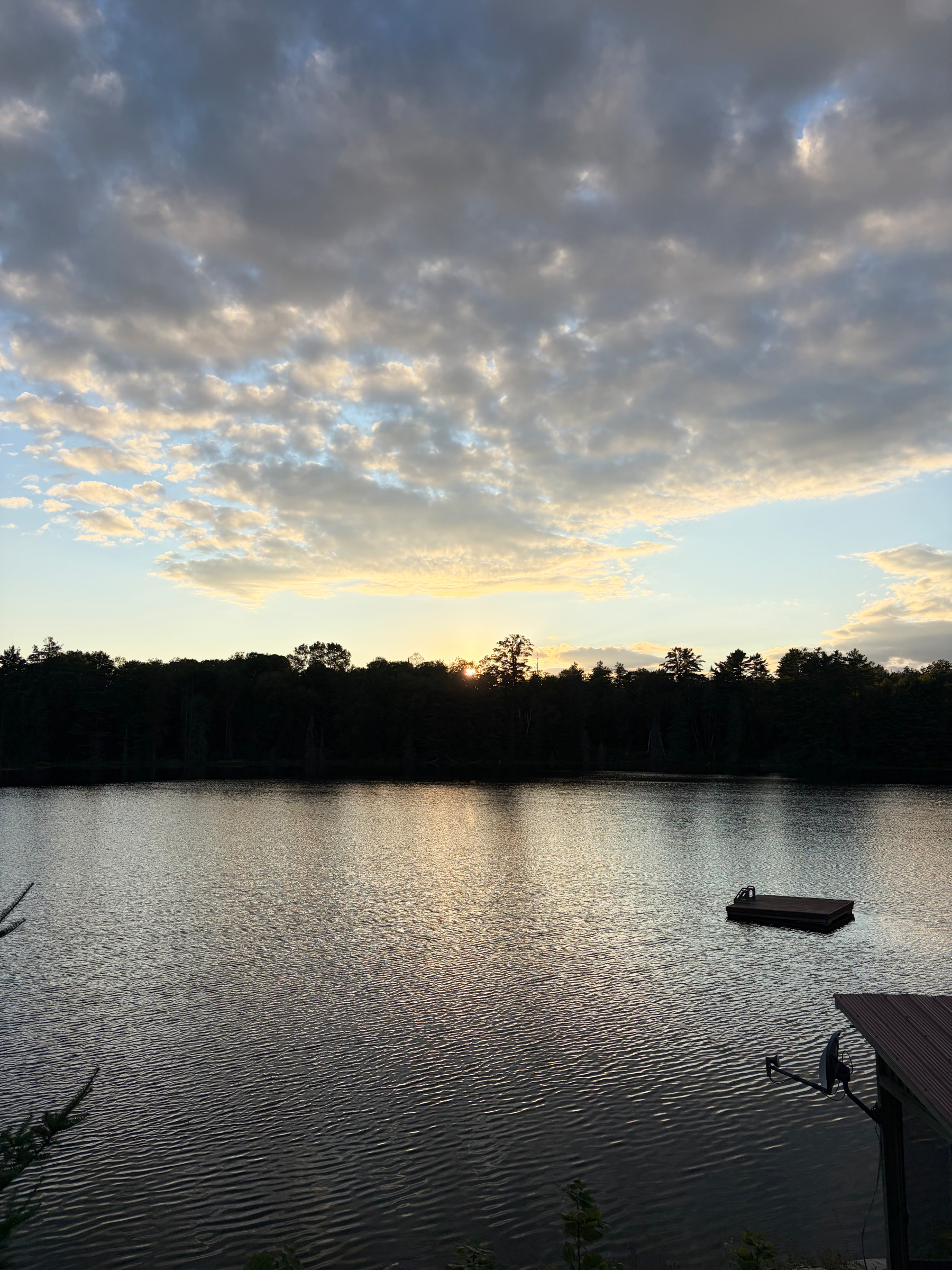 A serene lake reflecting the early evening sky with scattered clouds, bordered by a dark tree line and featuring a small floating dock.