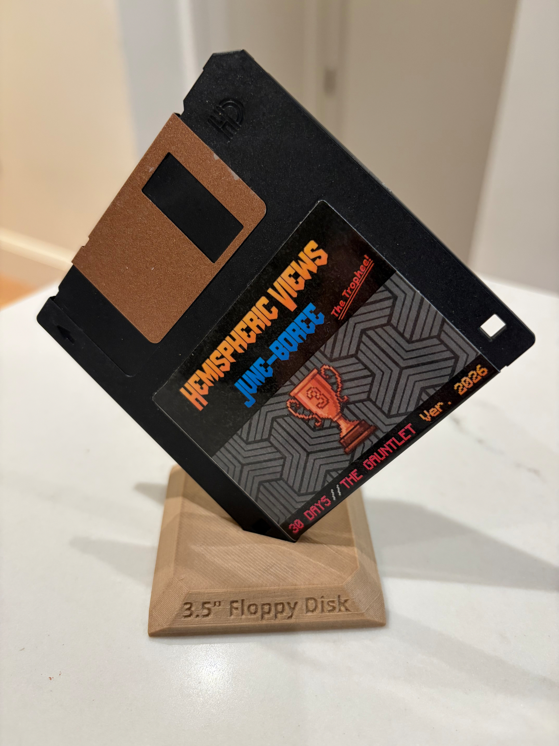 A 3.5 floppy disk labeled with Hemispheric Views, JUNE BOREE, and 39 DAYS / THE GAUNTLET Ver. 2026 is displayed standing on a wooden stand.