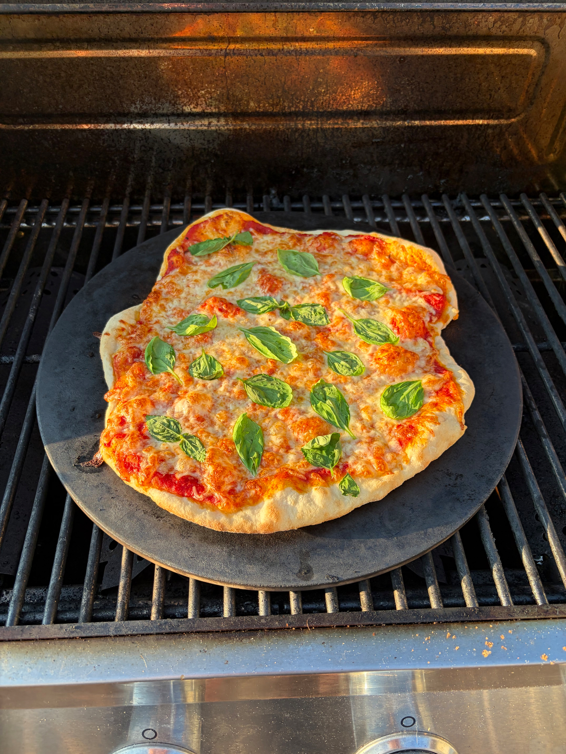 A homemade pizza topped with cheese and fresh basil leaves is baking on a pizza stone inside an oven.