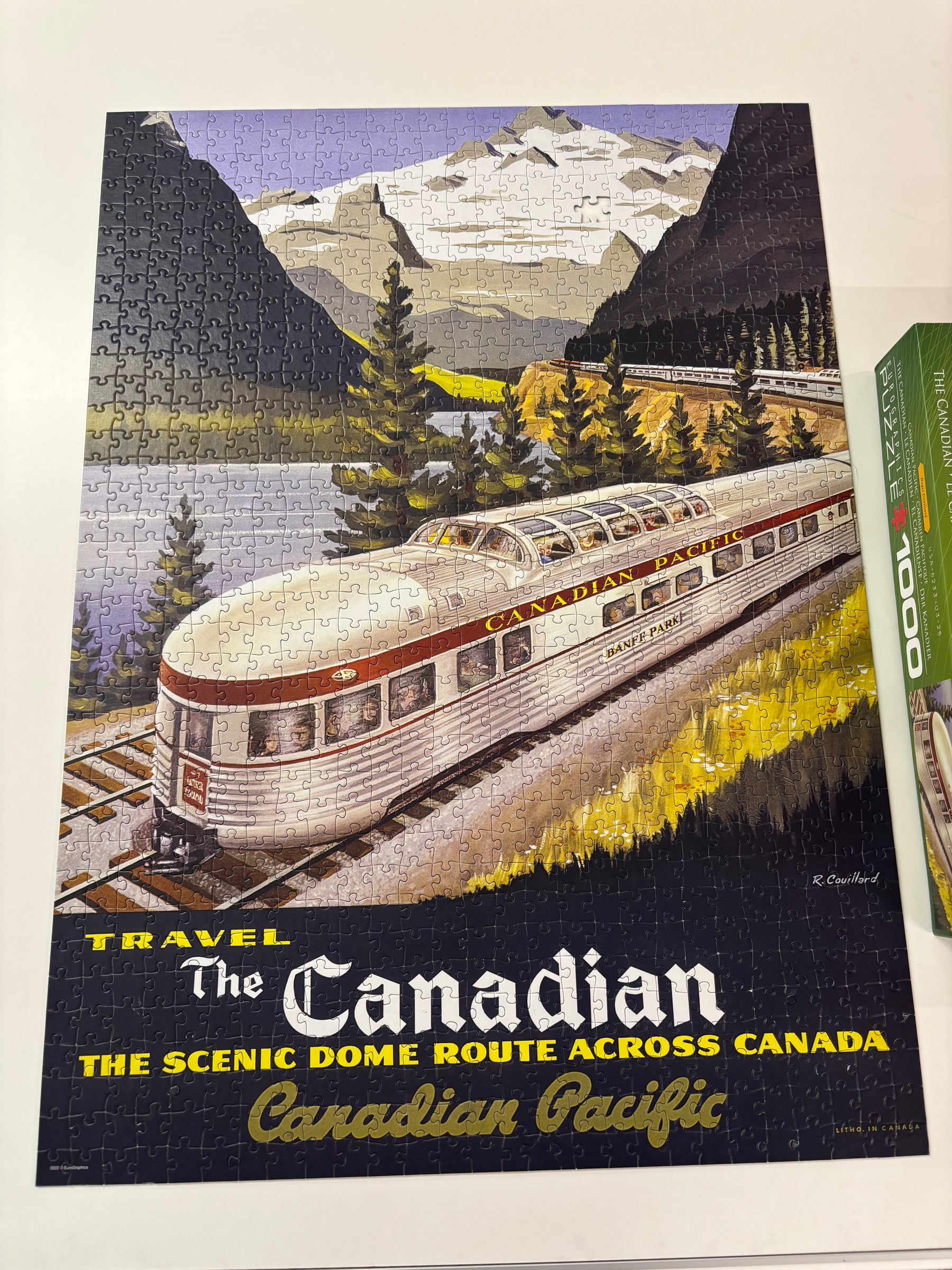 A completed puzzle depicts a vintage poster of The Canadian train traveling through a scenic landscape with mountains and forests, promoting a route across Canada.